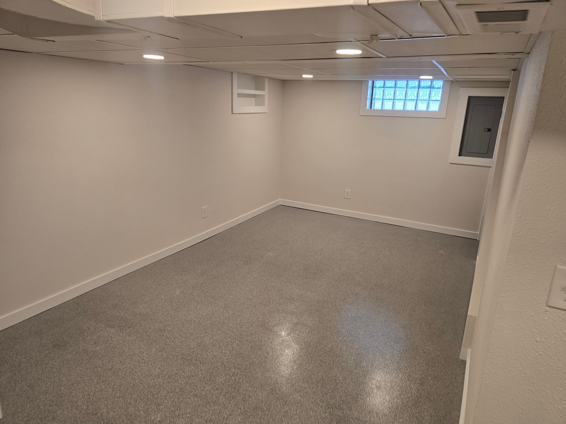 Empty basement room with gray carpet, white walls, drop ceiling, and a small window.