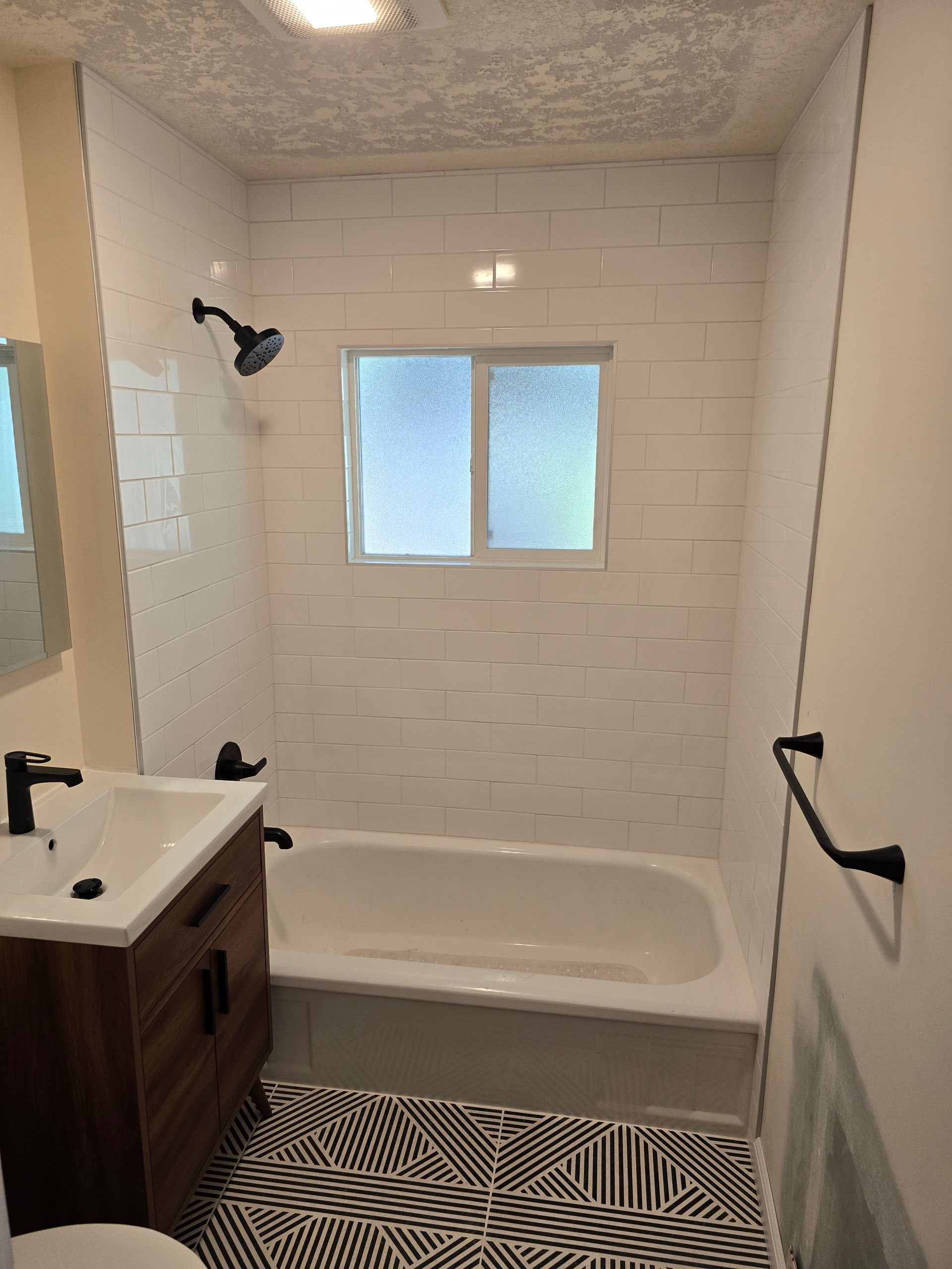 Small bathroom with a white bathtub, window, vanity sink, and black-and-white patterned rug.