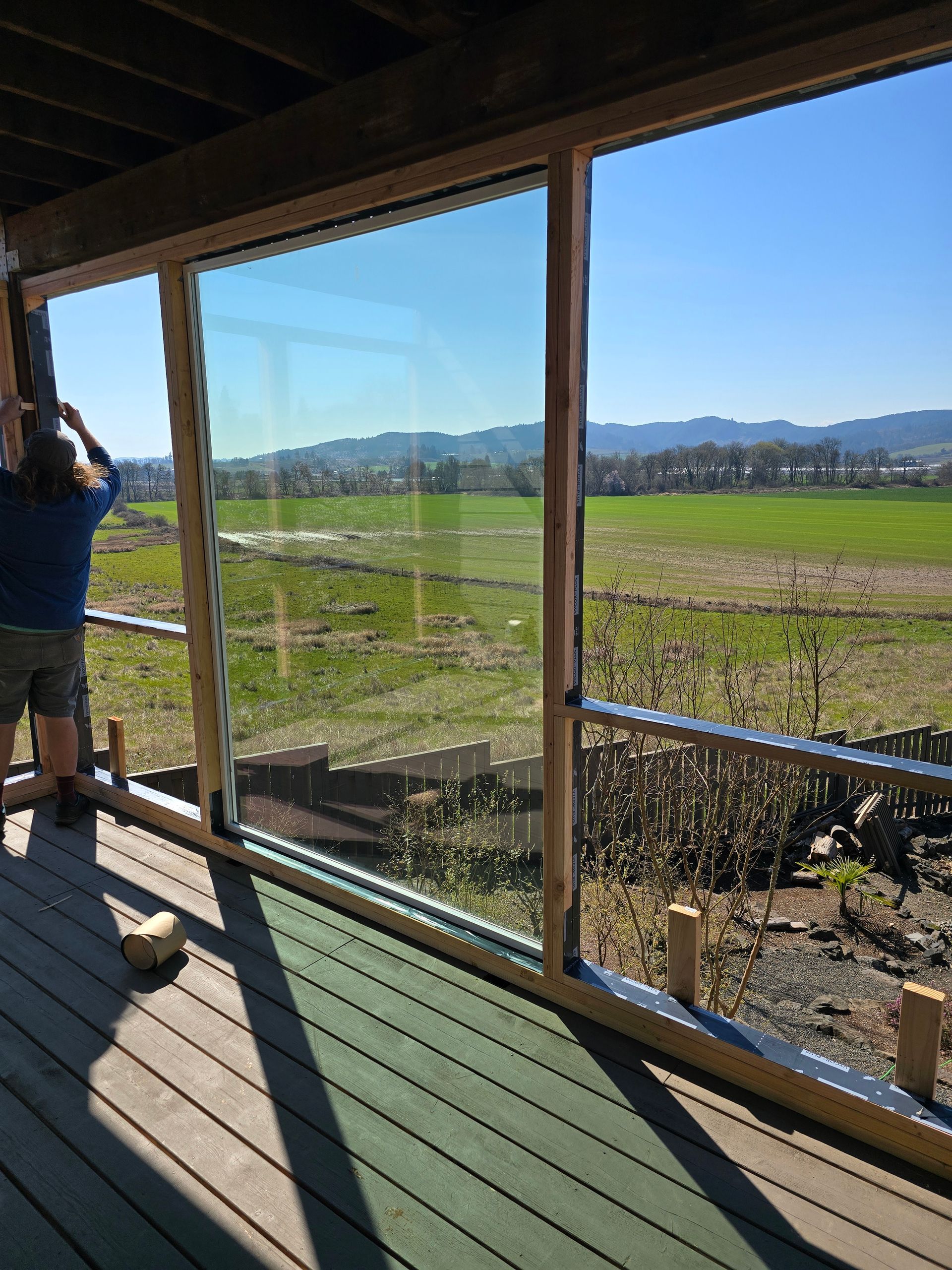 Person on a porch looks out over green fields and mountains through large windows.