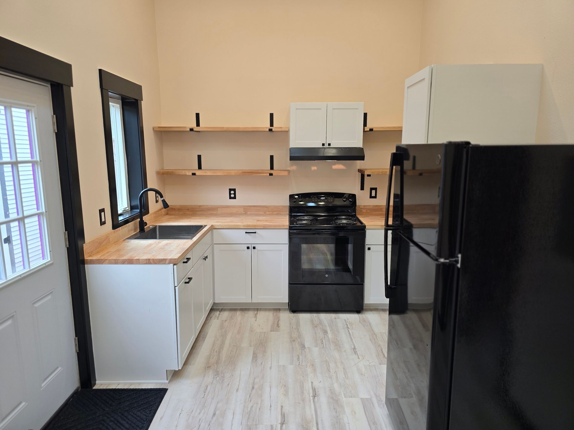 Bright kitchen with white cabinets, black appliances, light wood floor, and a door on the left.