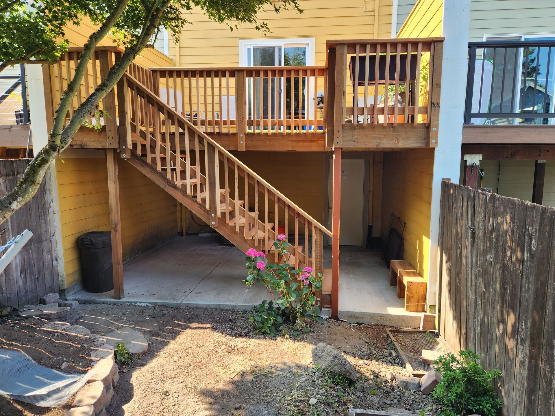 Backyard wooden deck with stairs leading to a yellow house, shaded patio below, and a small flower bed