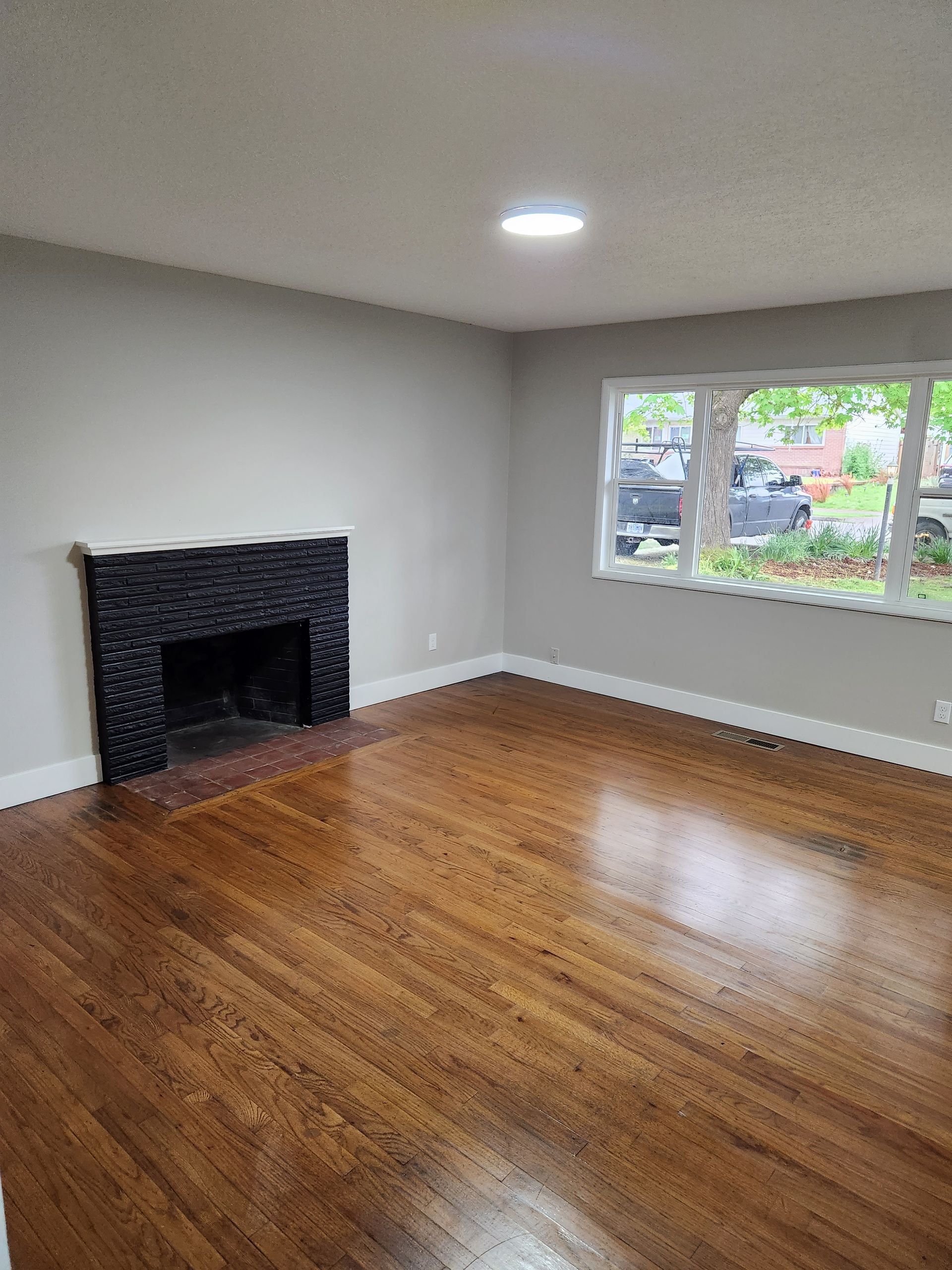 Empty living room with hardwood floors, a black fireplace, and a window overlooking greenery
