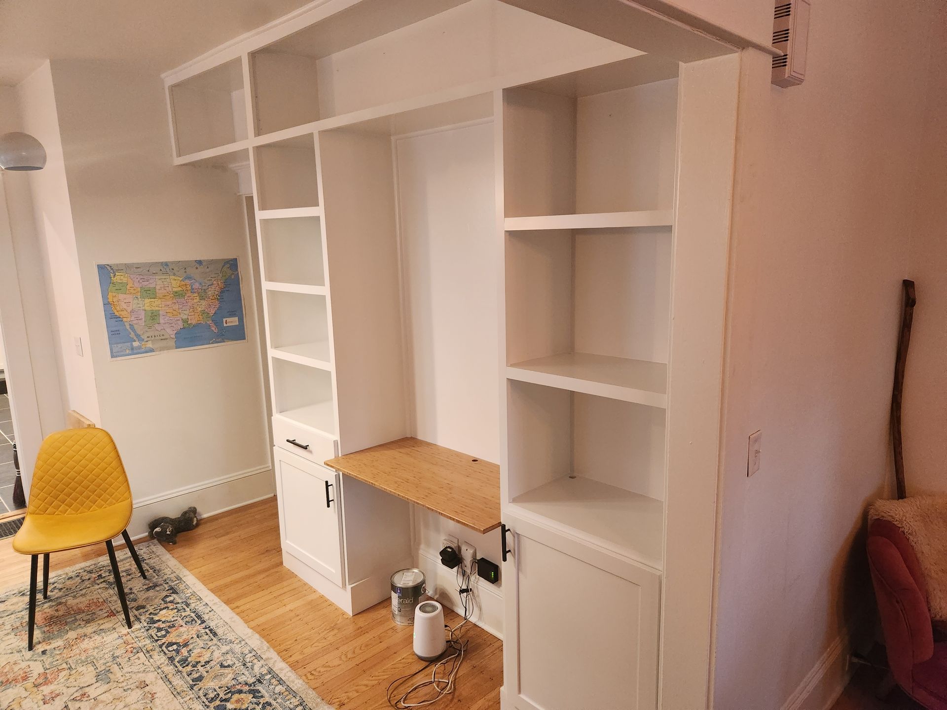 A white built-in desk and shelving unit in a small room, with a yellow chair and wooden floor.