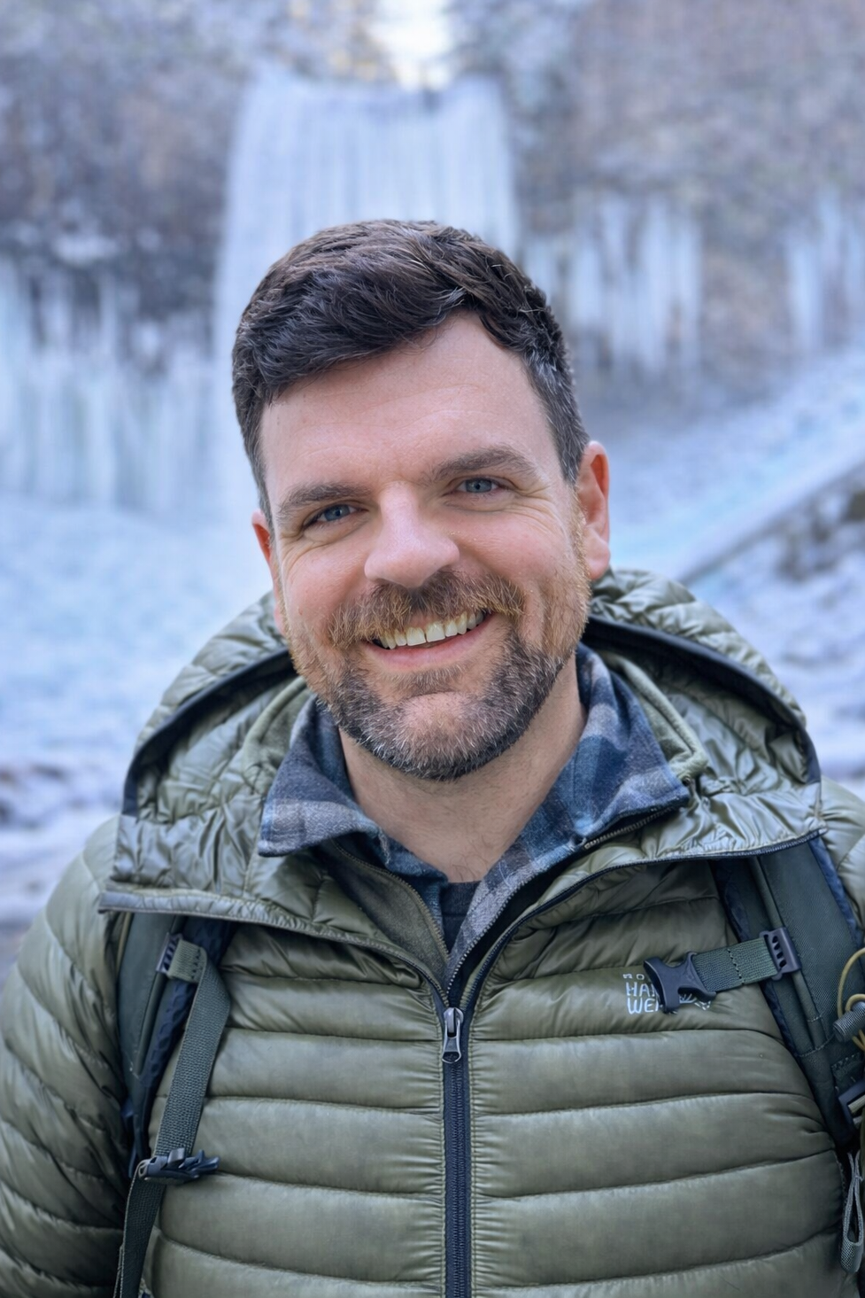 A smiling person in a green puffer jacket and backpack, posed in front of a partially frozen waterfall.