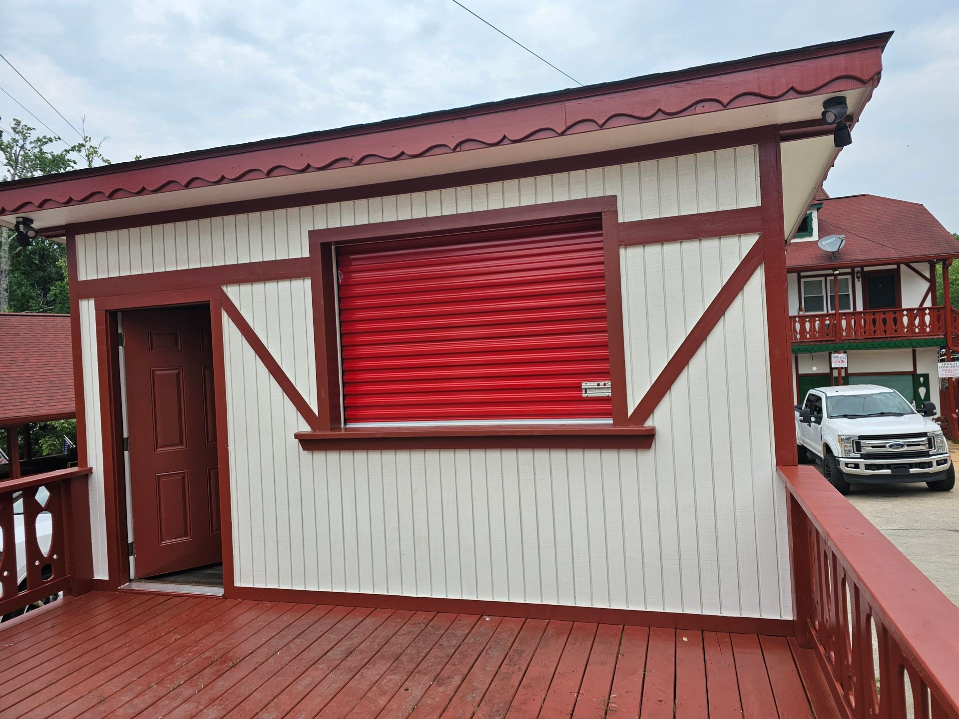 A white truck is parked in front of a small building with a red shutter on the window.