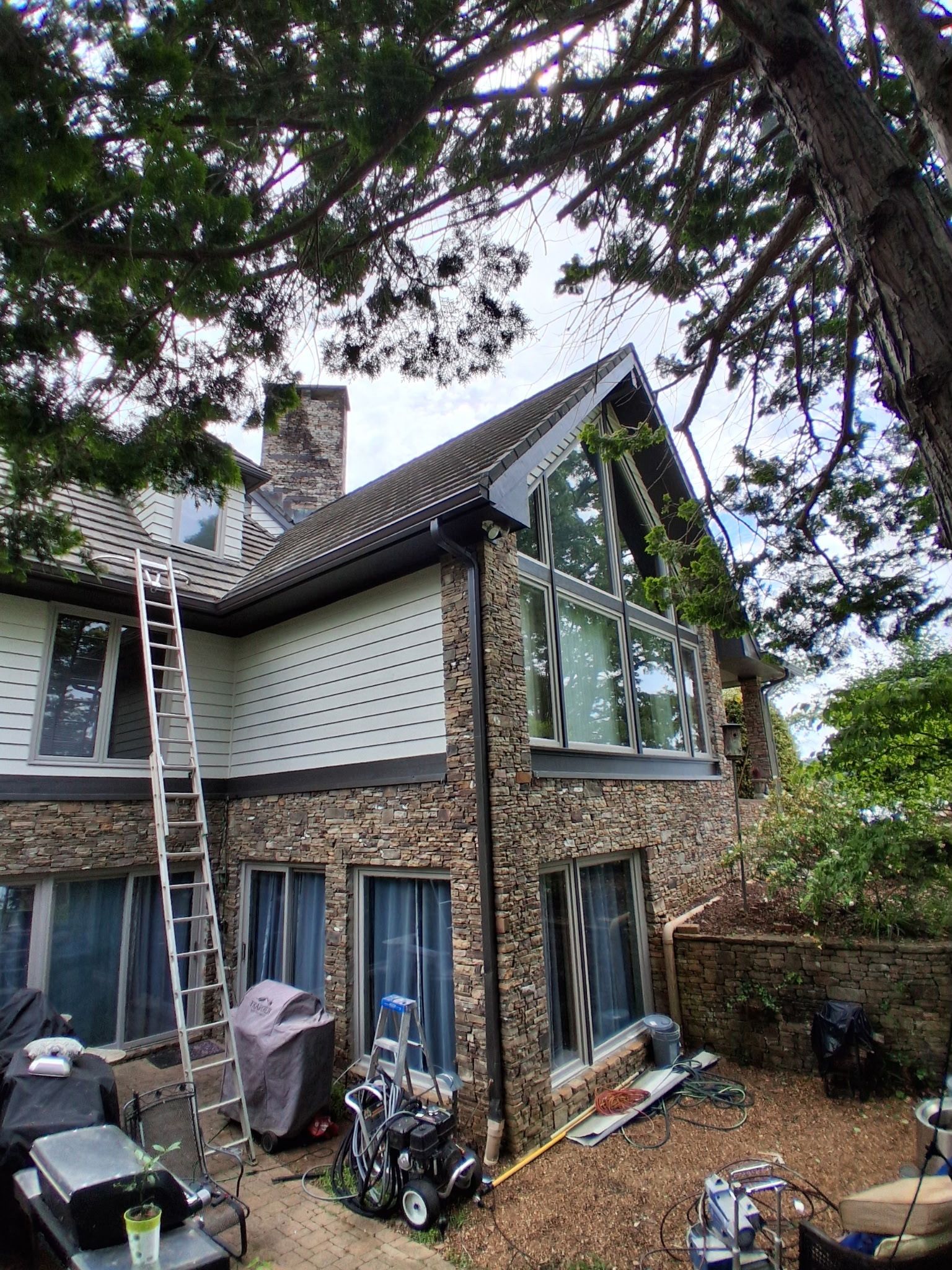Two-story house exterior with ladder, trees overhead, and yard equipment near the wall