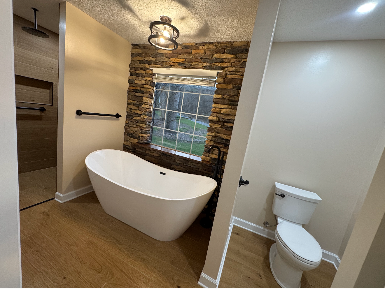 Modern bathroom with a freestanding tub, stone accent wall, window, and toilet on wood-look flooring.