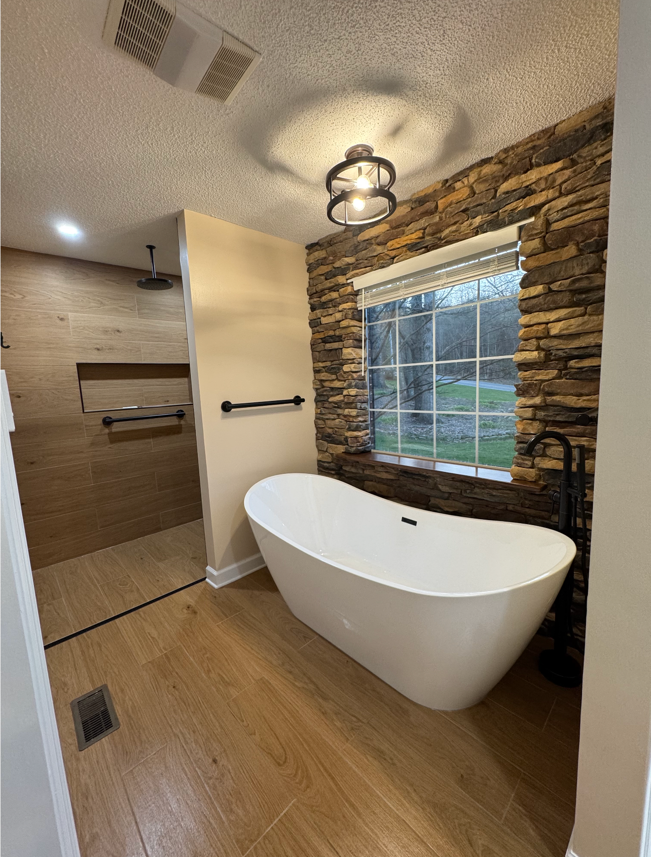 Modern bathroom with freestanding tub, stone accent wall, large window, and wood-look floor