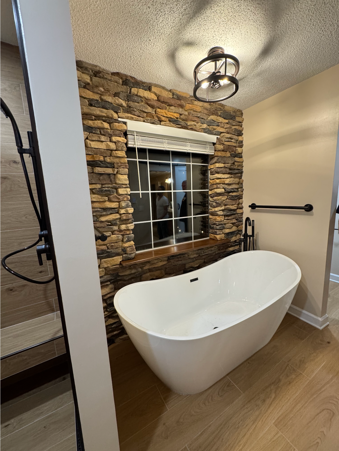 Modern bathroom with freestanding white tub, stone accent wall, window, and wood floor
