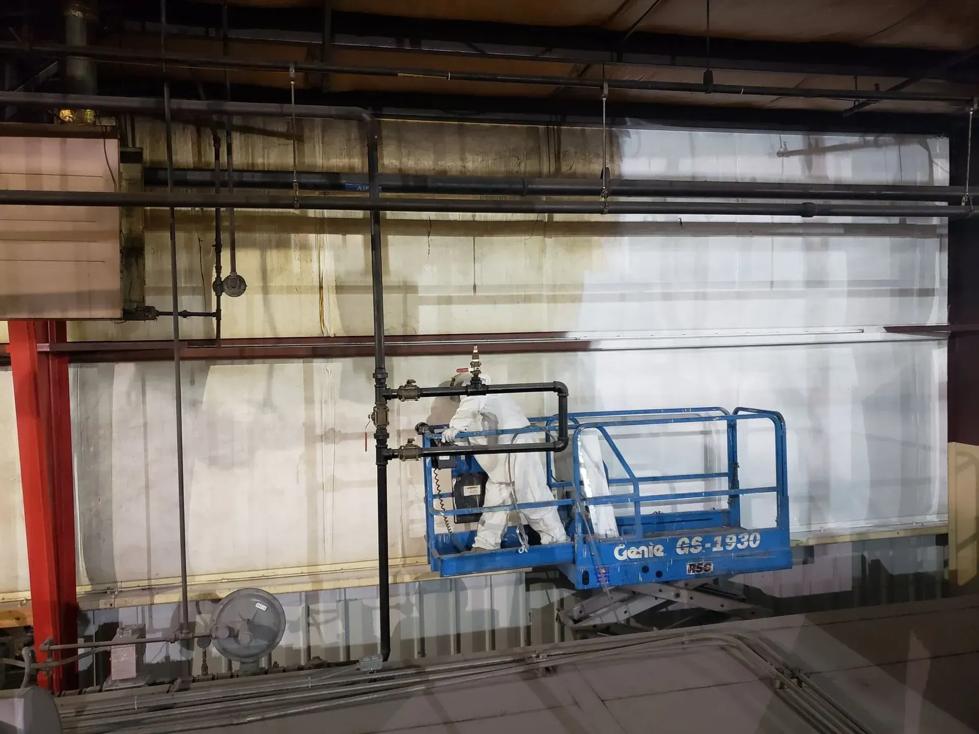 A person in a blue lift working on a white wall inside a building with pipes.