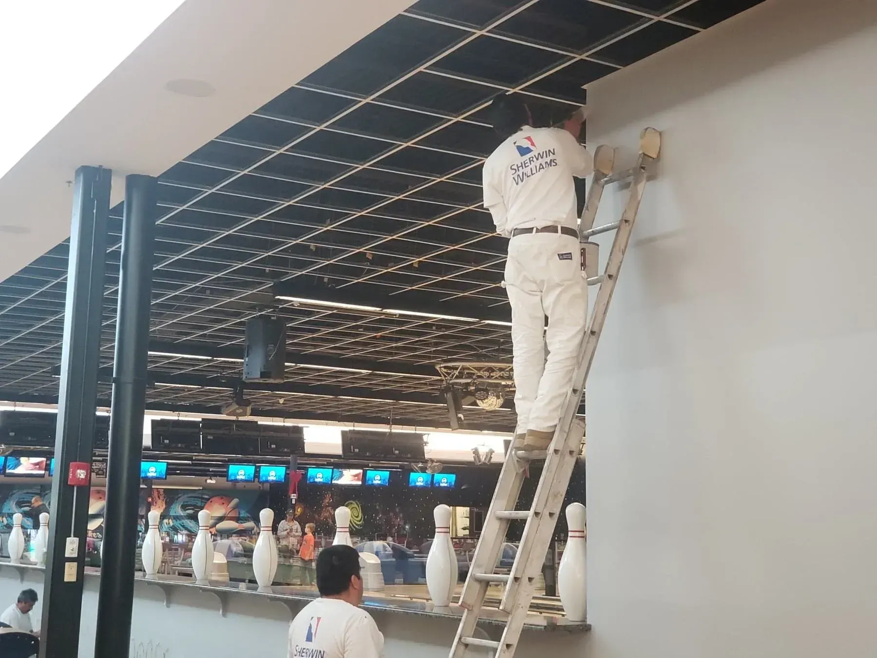 Painter in white overalls on a ladder, painting a light grey wall in a bowling alley.