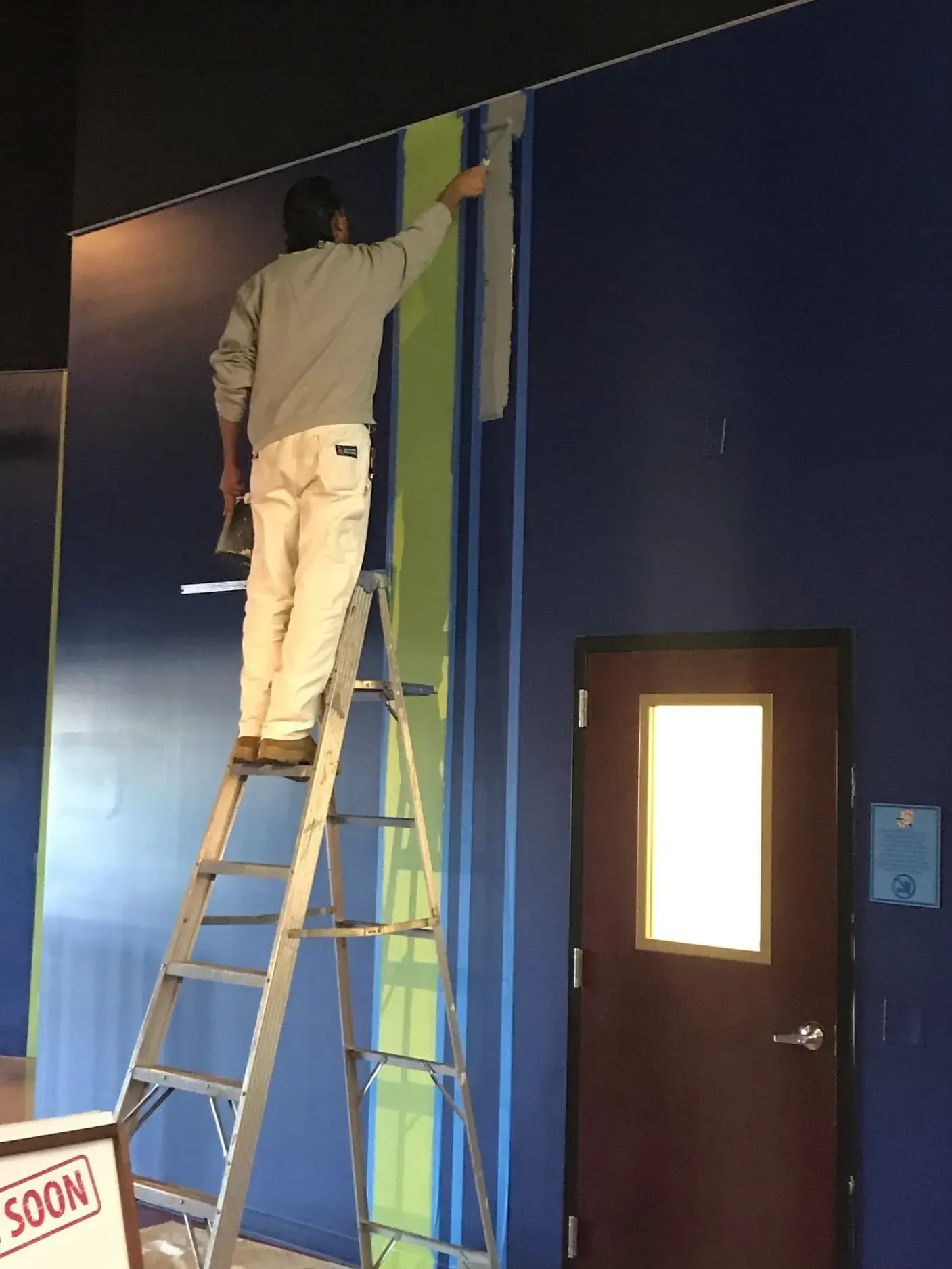 Person painting a blue wall while standing on a ladder. They are in front of a door.