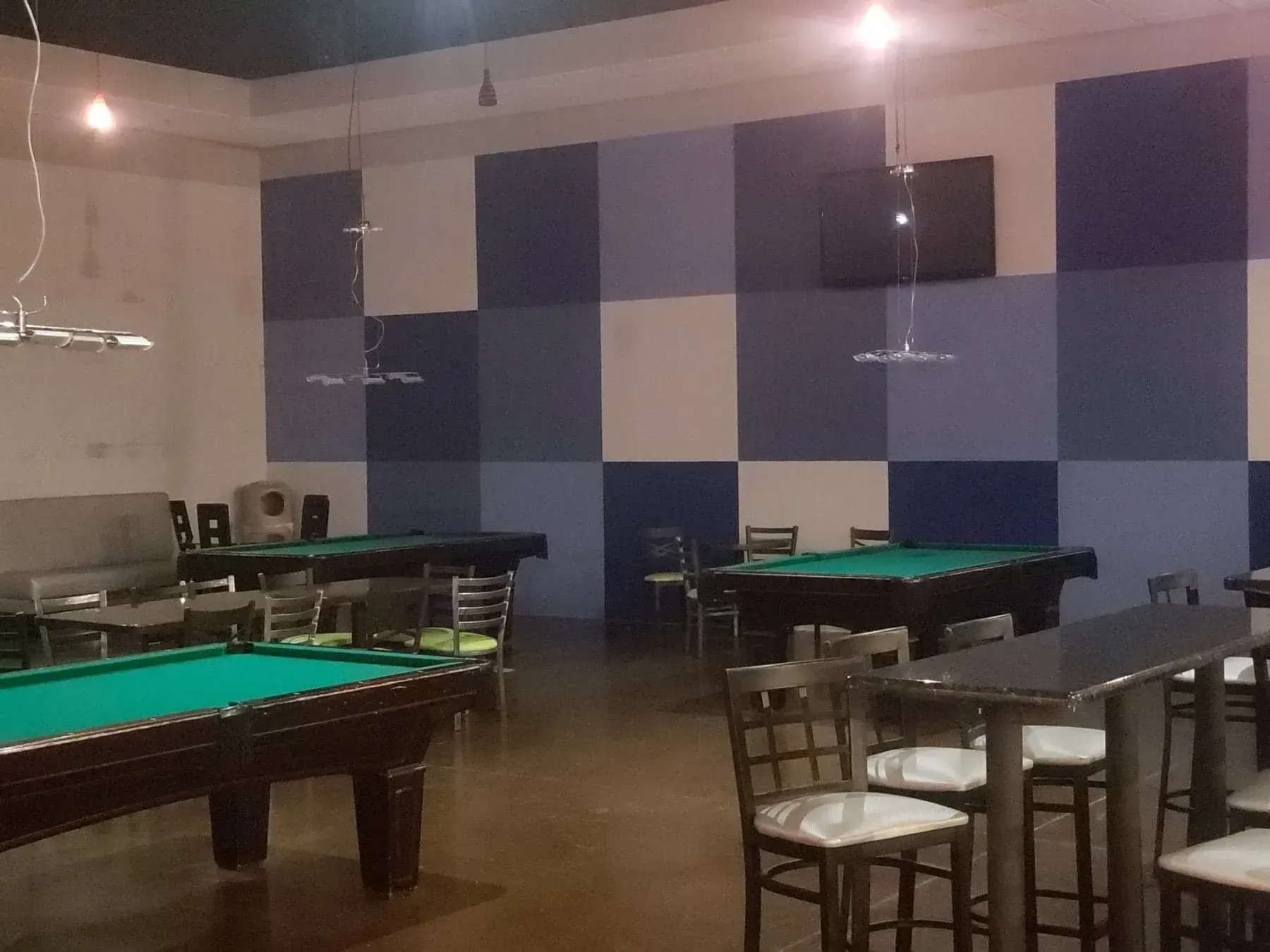 A pool hall with three green-felt tables, blue and white square wall design, and seating.