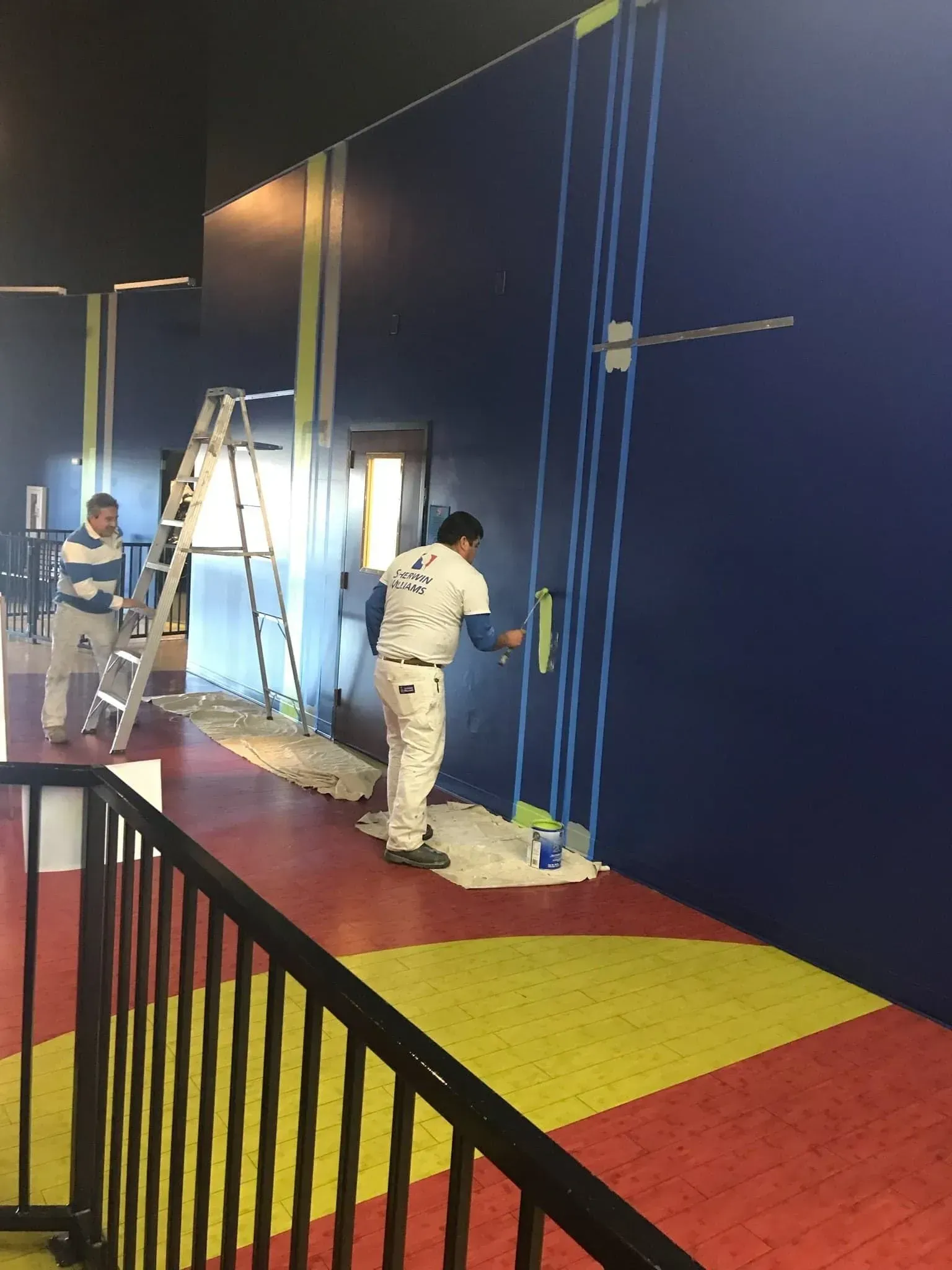 Two painters applying blue paint to a wall. One on a ladder, the other using a roller. Interior setting, color-blocked floor.