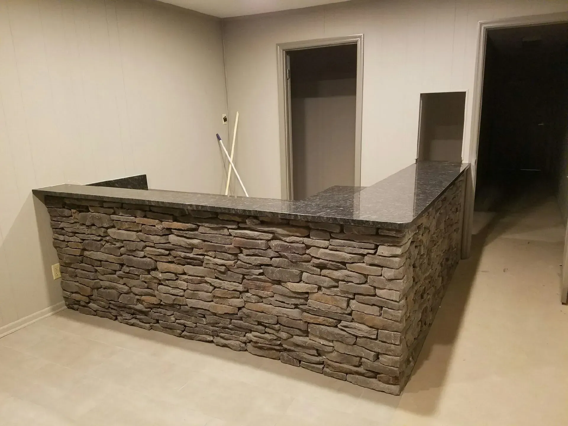 A stone-faced bar with a dark countertop in a room with neutral-colored walls and flooring.