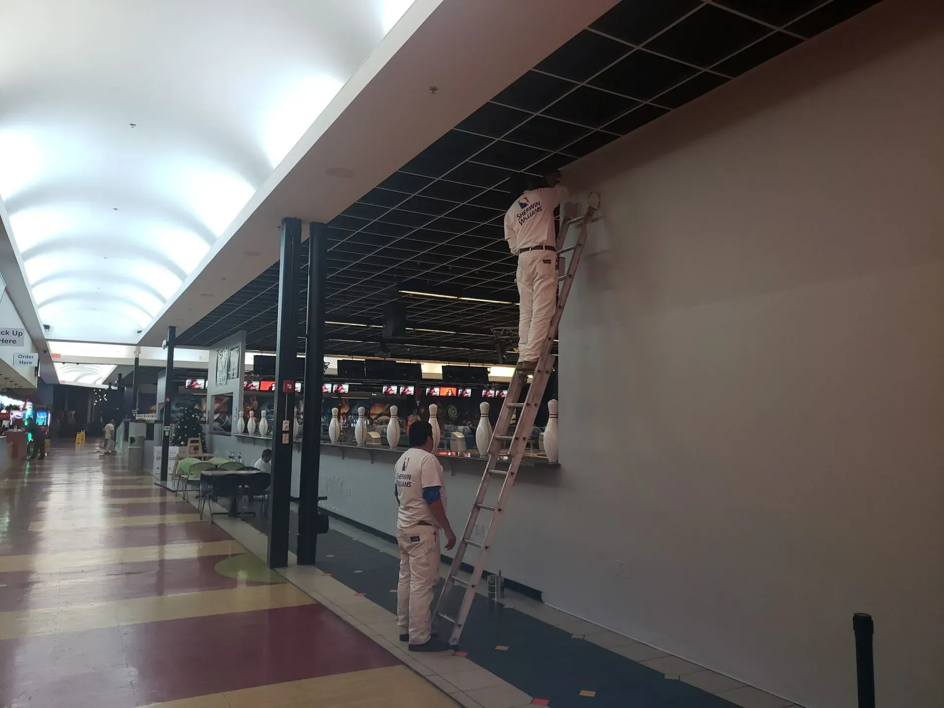 Two painters in white coveralls painting a gray wall in a shopping mall. One is on a ladder.