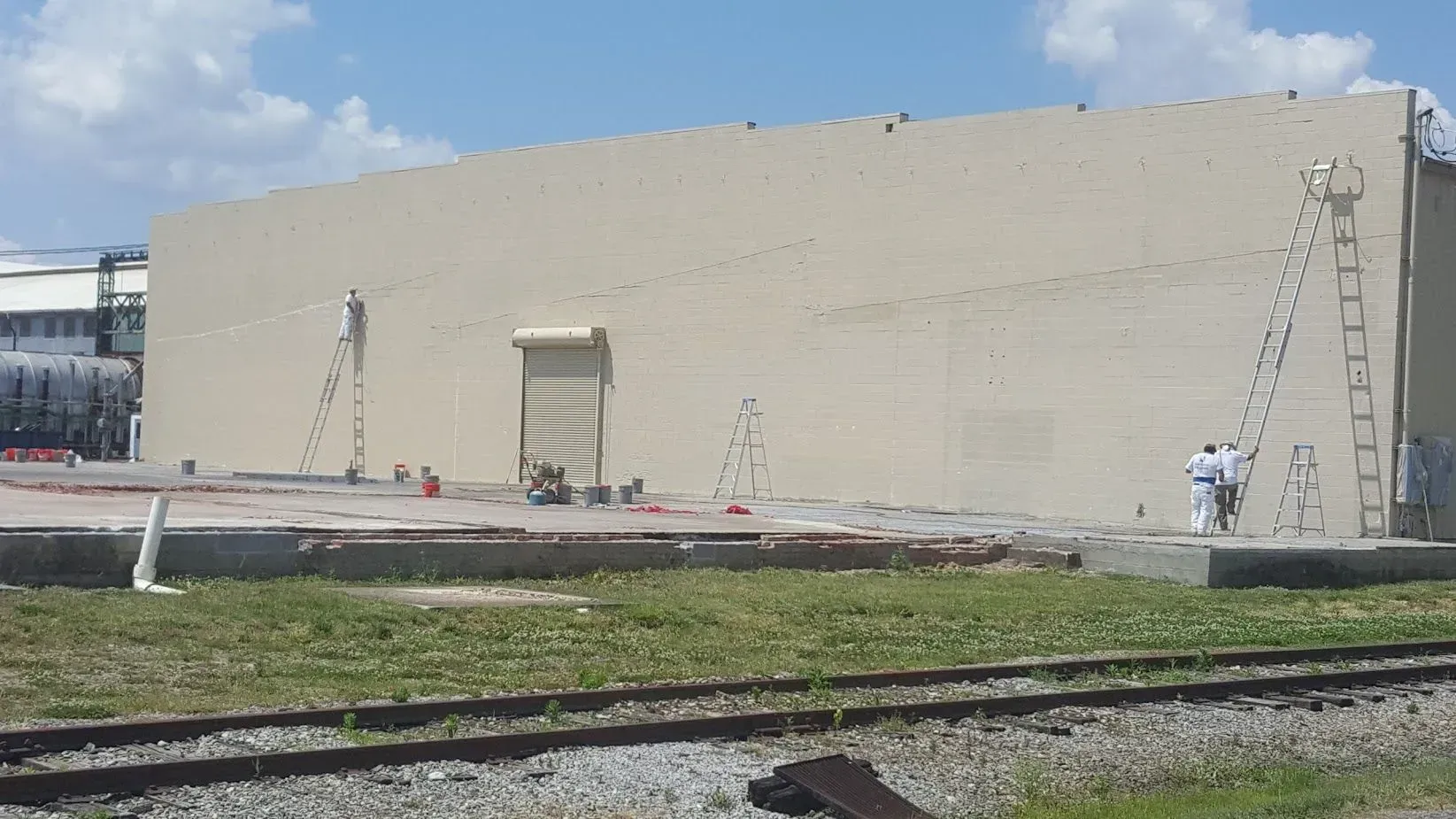 Exterior of a beige industrial building, ladders on the wall, railroad tracks in the foreground.