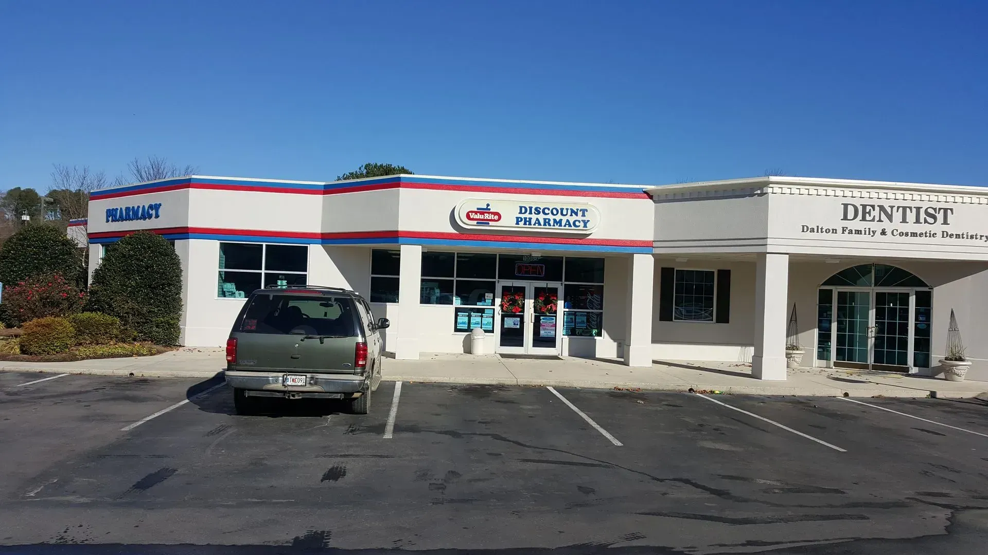 Exterior of a pharmacy and dentist office in a strip mall, with a car parked out front on a sunny day.