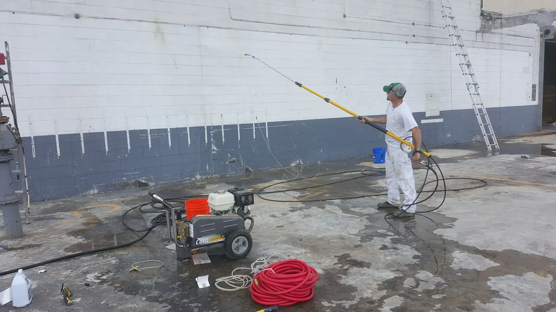 A person spray paints a building's exterior with a long handled sprayer.