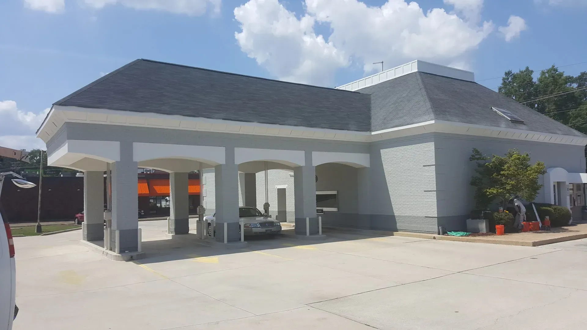Gray bank building with drive-through lanes under a covered area; parked car visible. Sunny day.