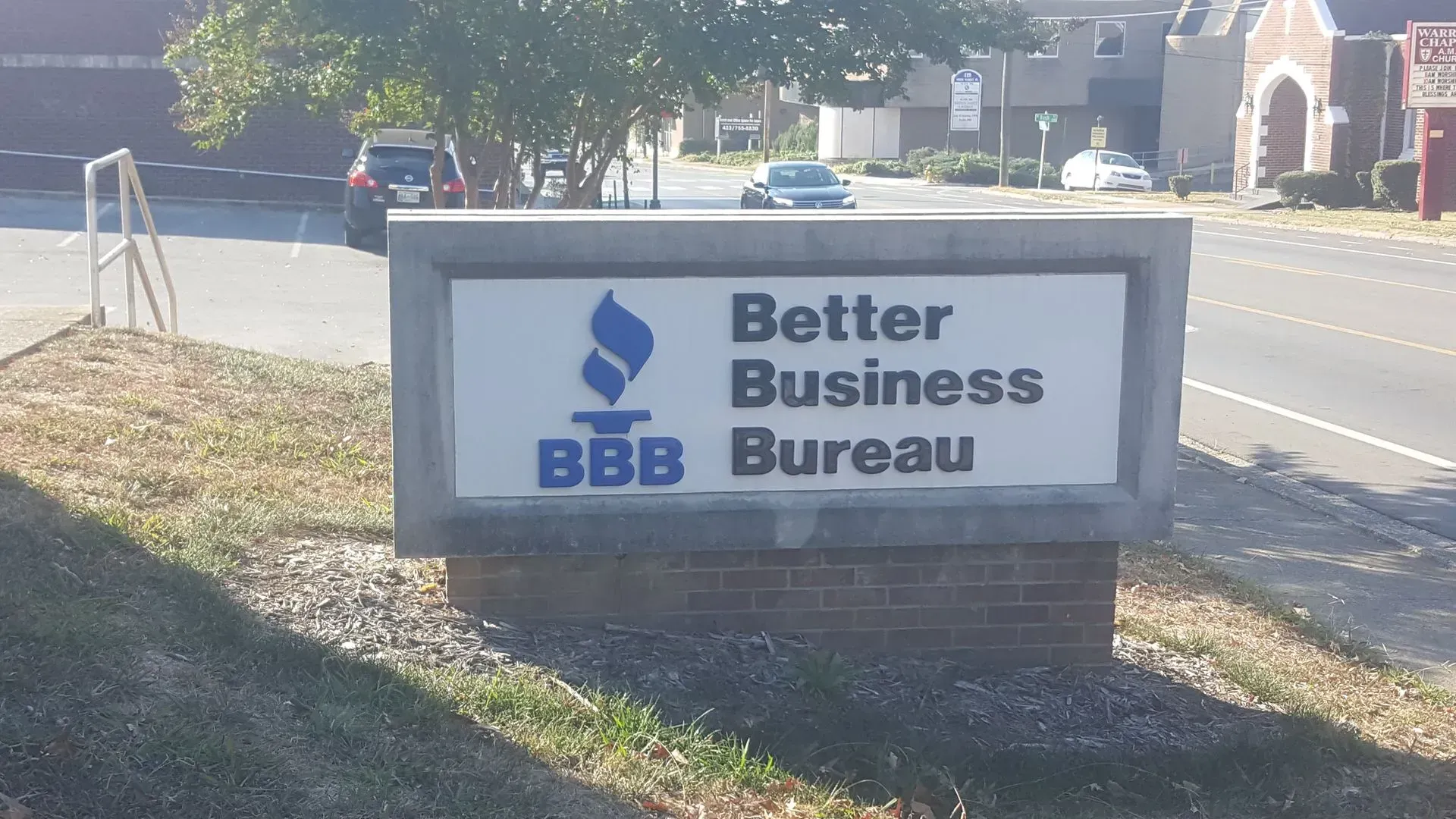 Sign for the Better Business Bureau, featuring their logo, set outdoors on a brick base.