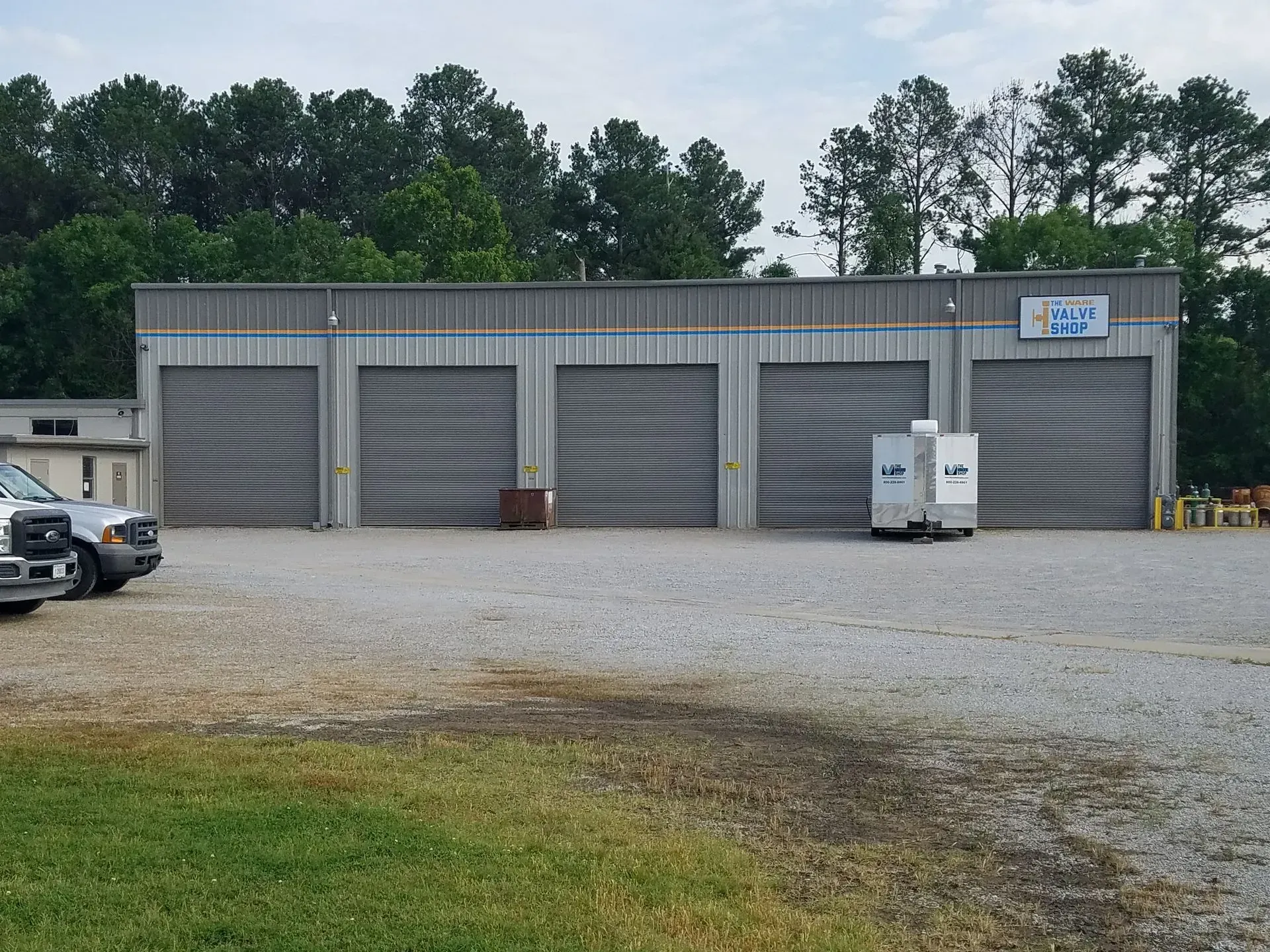A storage facility with four garage doors; gravel lot, with truck and greenery in the background.