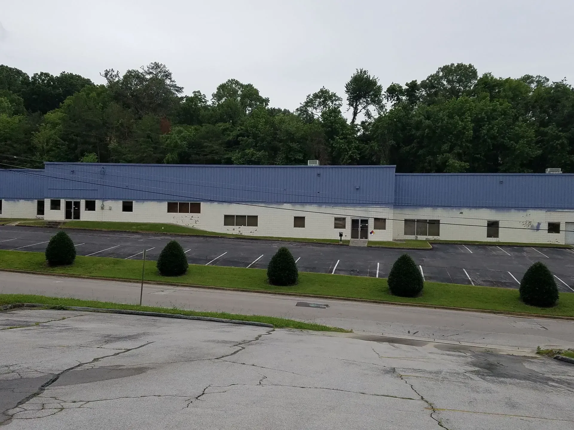 A long, one-story building with a blue roof, in front of a treeline, and a cracked parking lot.