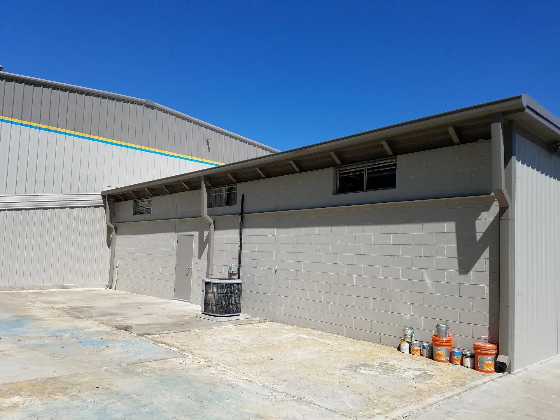 A gray building with small windows and a brown roof, next to a larger metal building, under a blue sky.