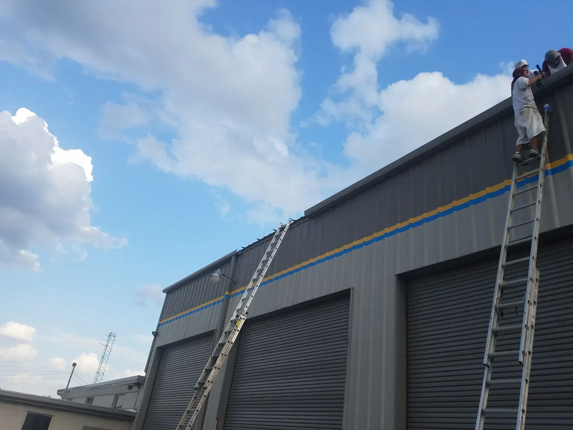 Workers on ladders painting a gray industrial building with a blue and yellow stripe under a cloudy sky.