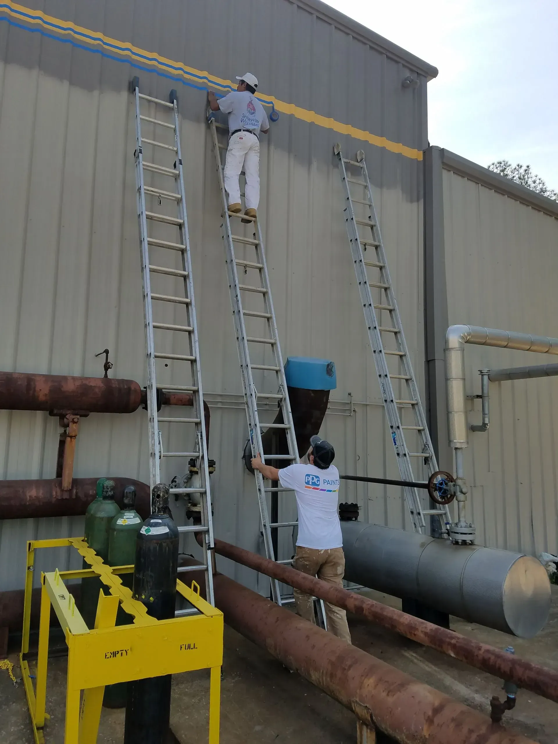 Two workers on ladders painting a building's exterior. One at top, one mid-level, near pipes and equipment.