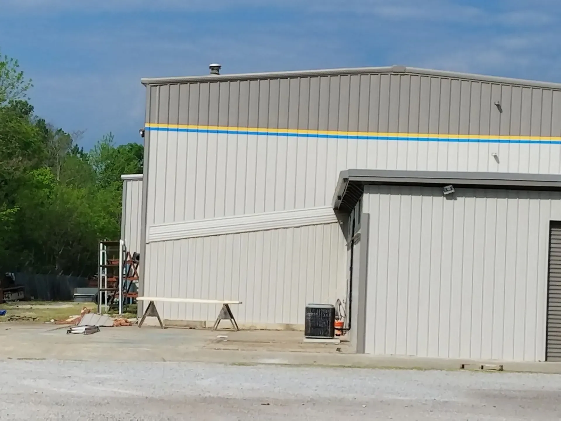 Warehouse exterior with white metal siding, blue and yellow trim, and gravel parking area.