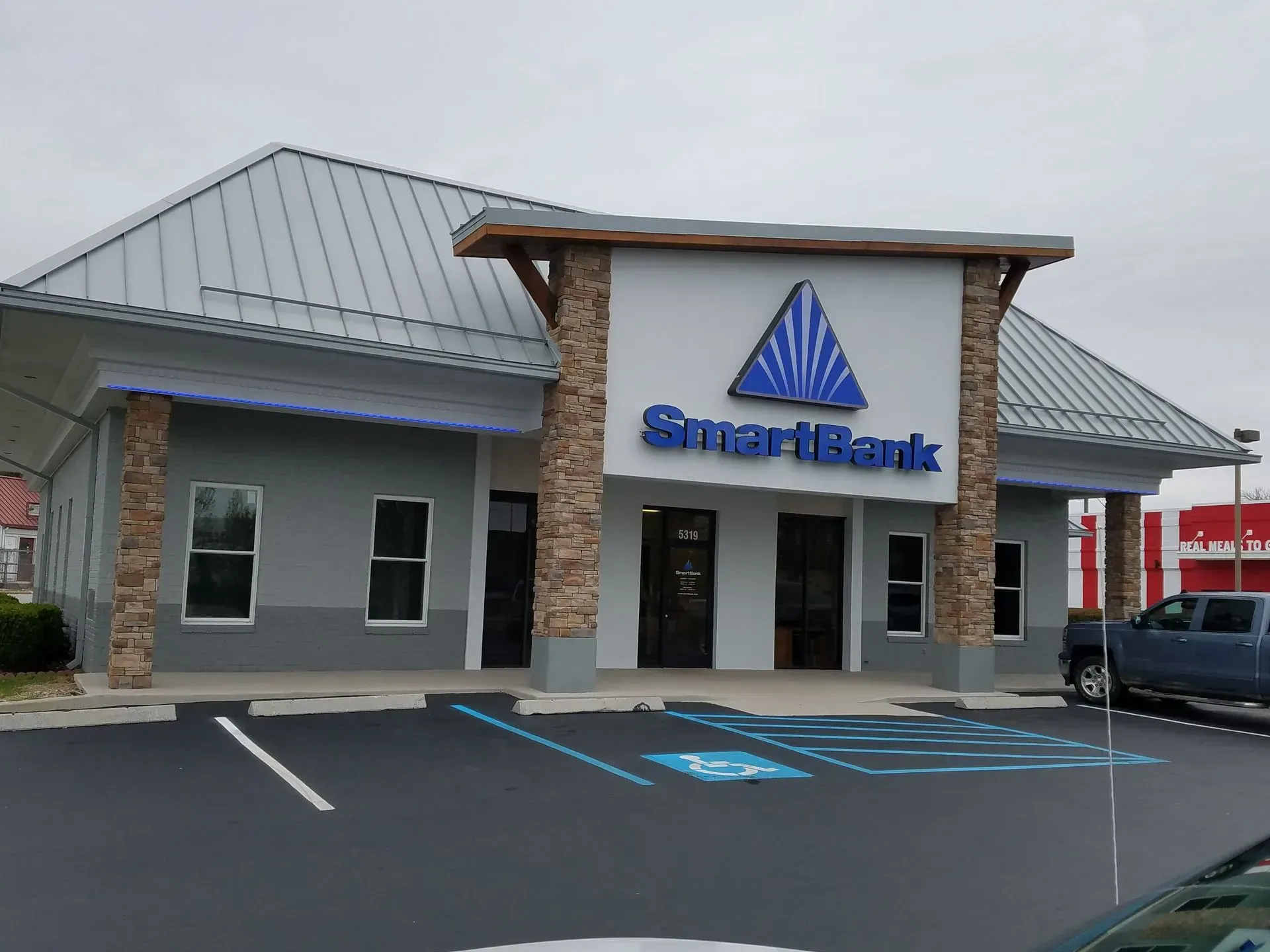A SmartBank building with a blue logo, gray exterior, and stonework pillars, with a cloudy sky.