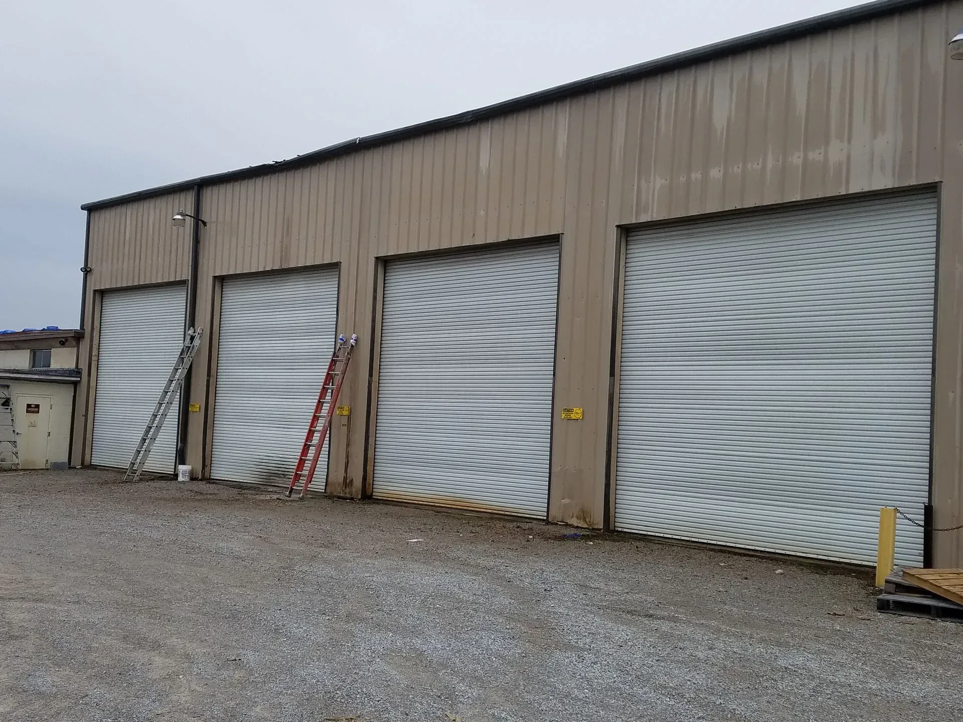 Three garage doors on a beige metal building. A ladder is leaning against one door. Gray gravel.