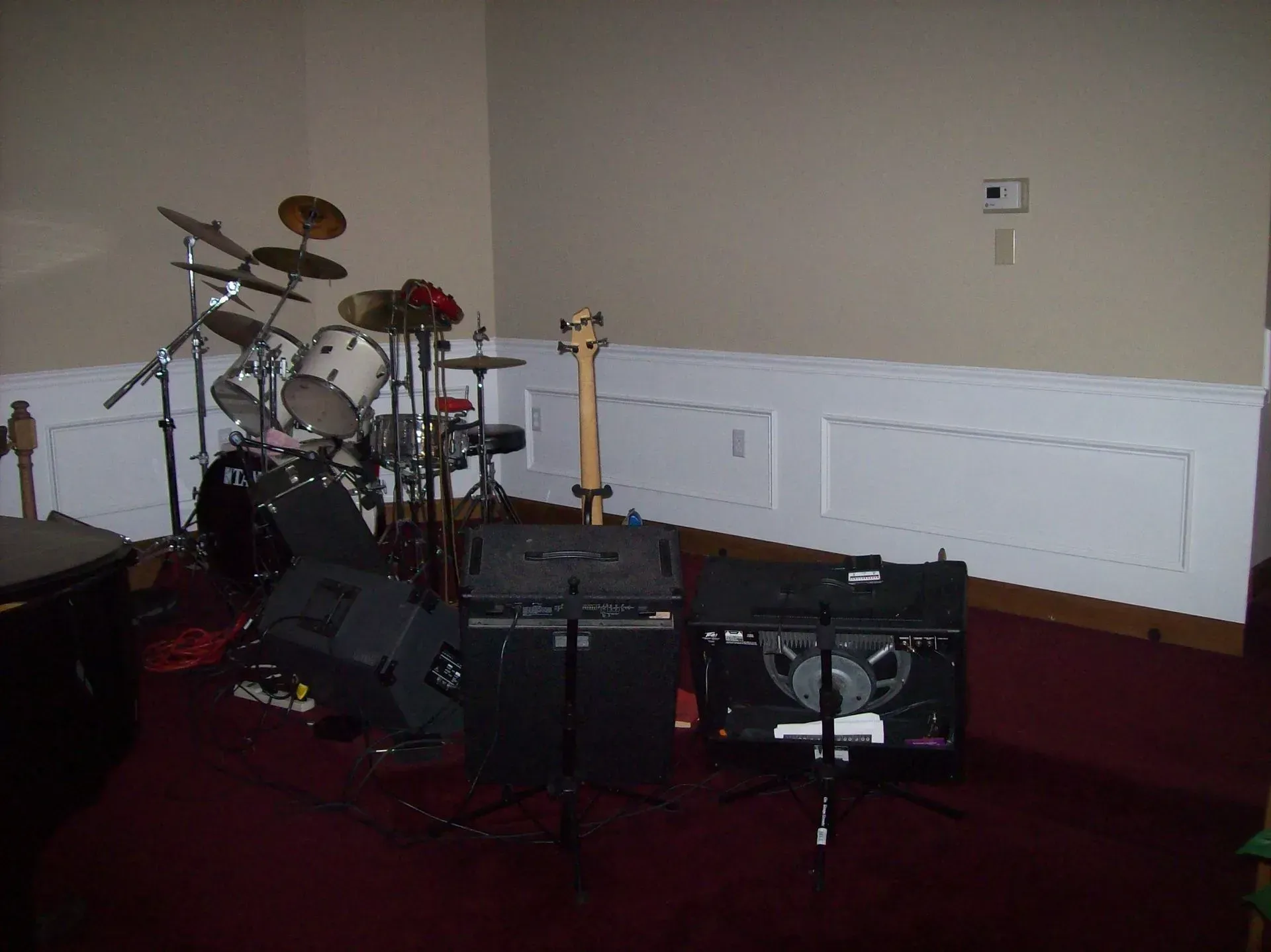 A drum set, bass guitar, and amplifiers set up in a room with red carpet and white wainscoting.