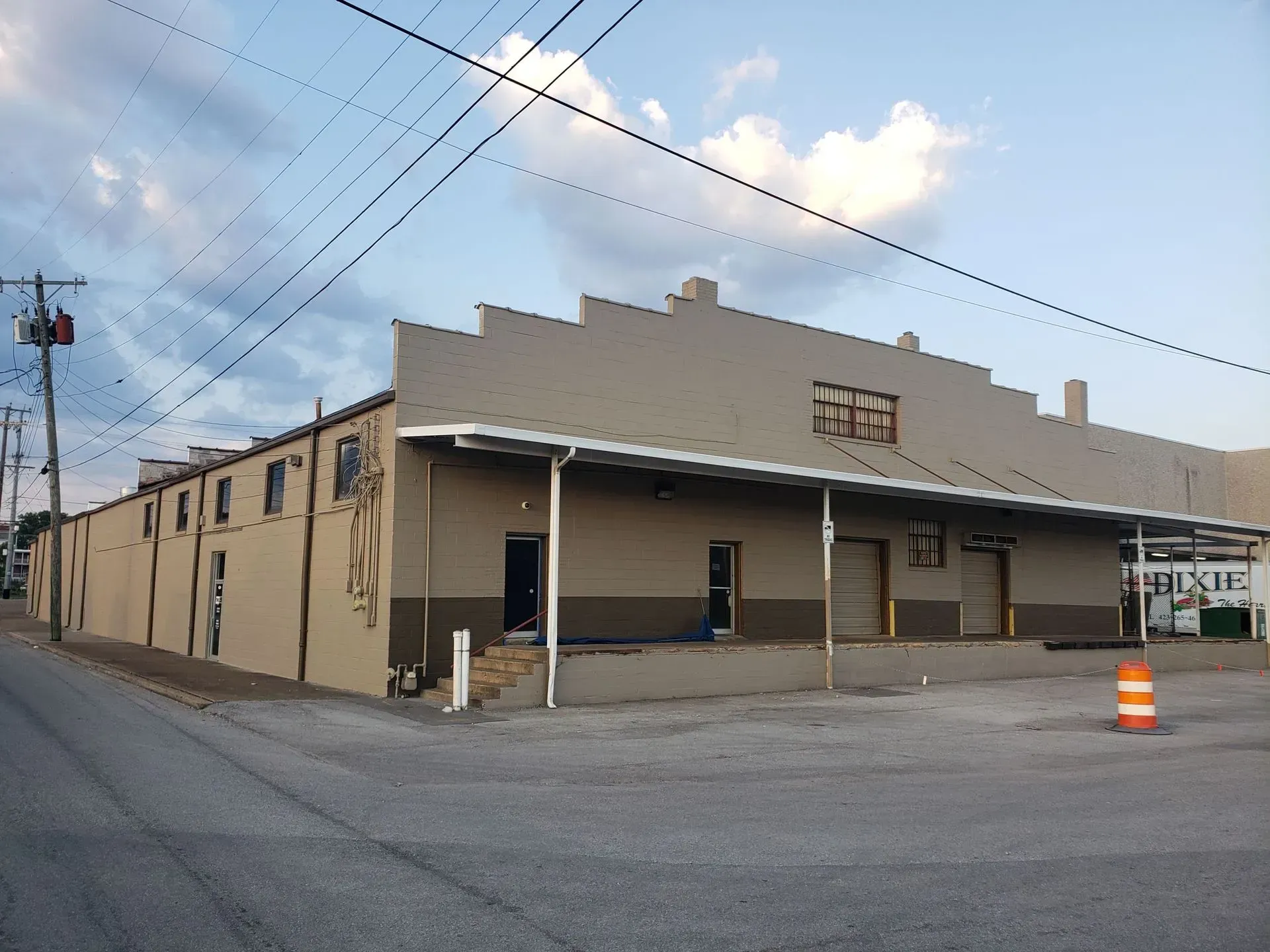 Tan industrial building with angled roofline, loading docks, and power lines against a cloudy sky.