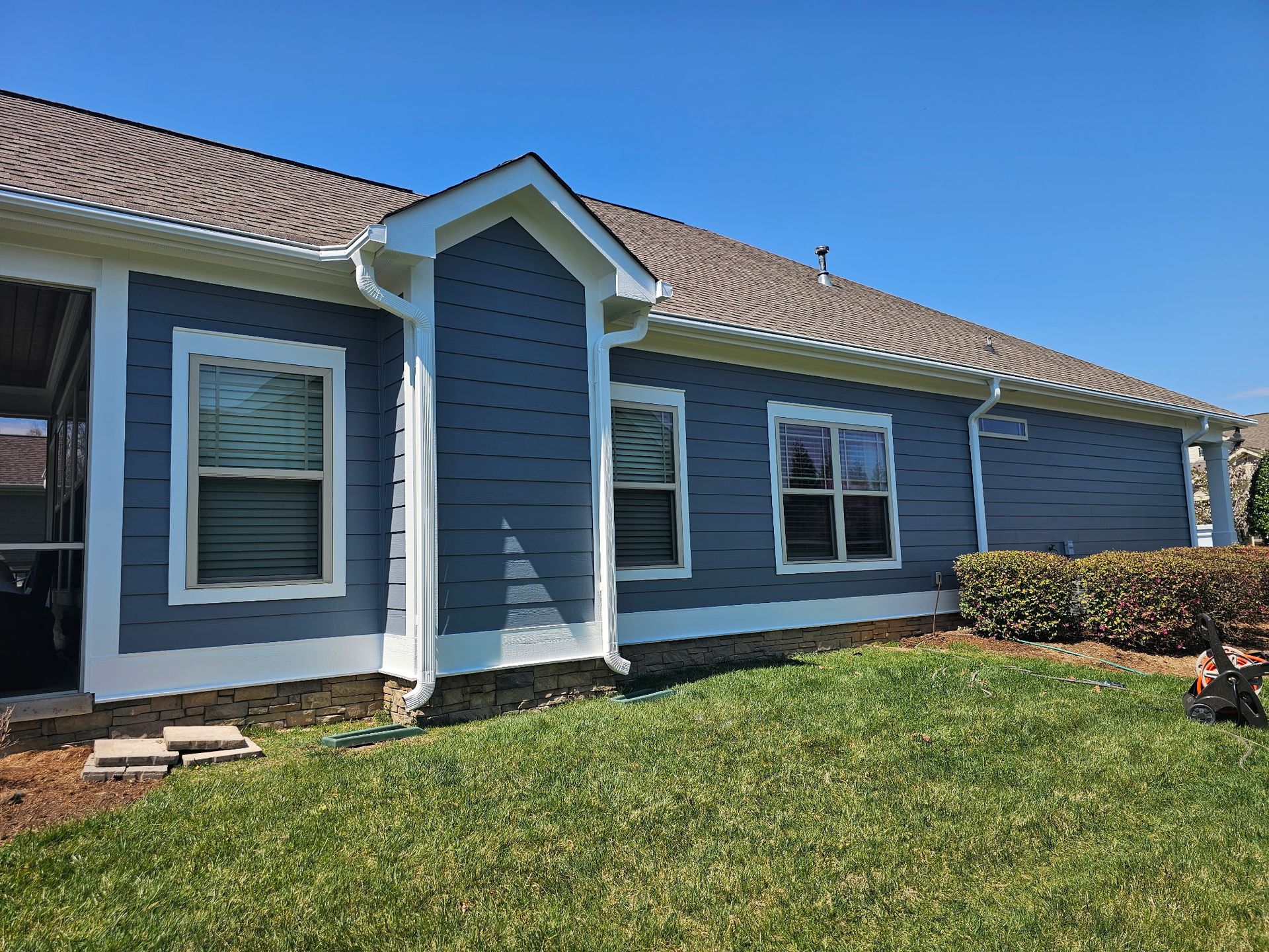 Blue-sided house with white trim and gutters, brown roof, windows, and green grass on a sunny day.