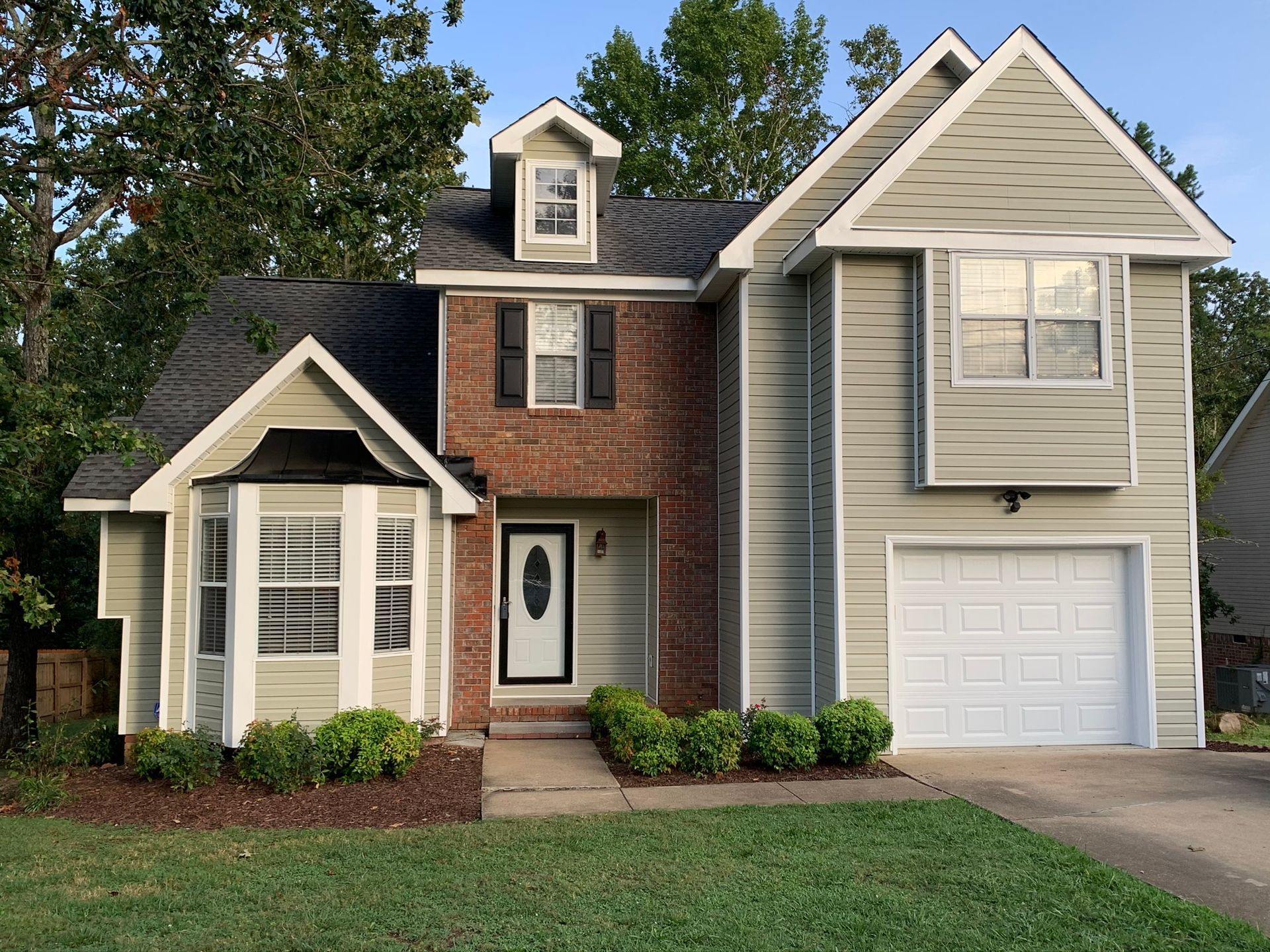 Two-story house with gray siding, a brick facade, and a white garage door. Green lawn and bushes.