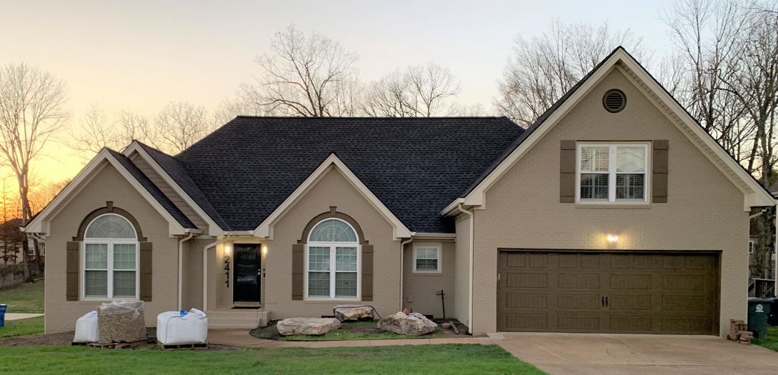Tan stucco house with brown garage door and shutters. Trees and sunset in the background.