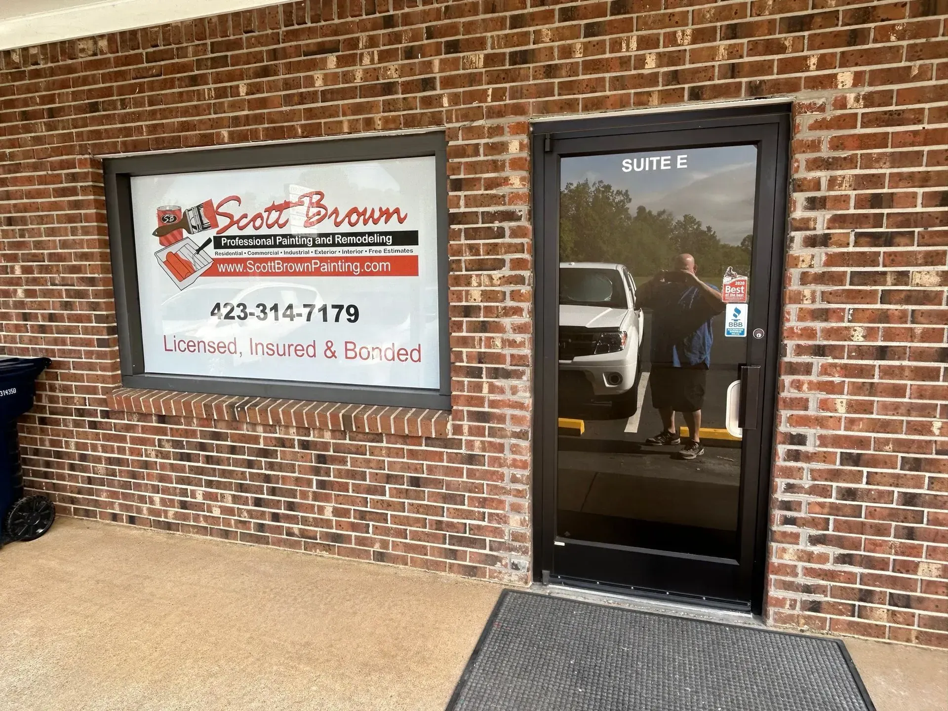 Exterior of Scott Brown Plumbing office with window sign and door.  Brick facade.  White truck visible through the door.