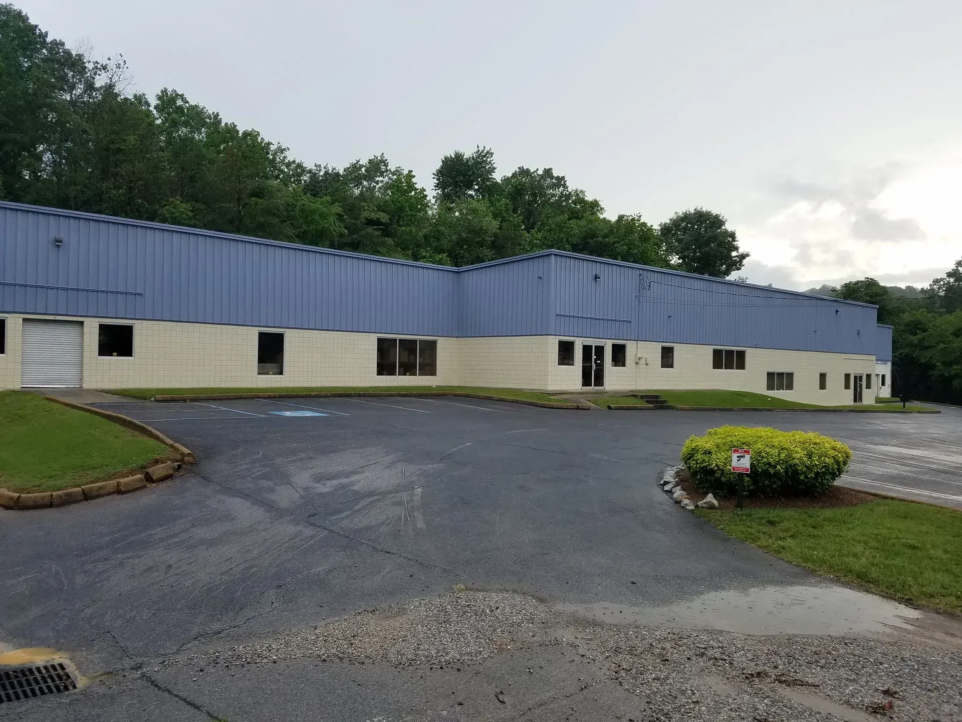 Gray, wet asphalt leads to a light-yellow industrial building with a blue roof and green trees.