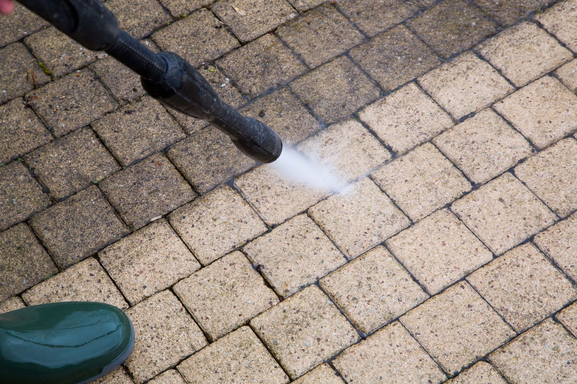 A pressure washer cleaning a brick patio; half cleaned, half dirty; green boot in view.