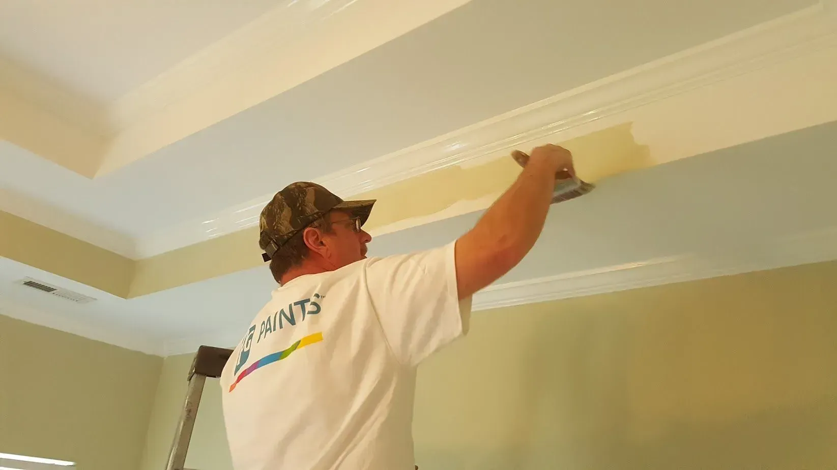 Man on ladder, painting wall trim with a putty knife. Interior room with tan walls and white ceiling.
