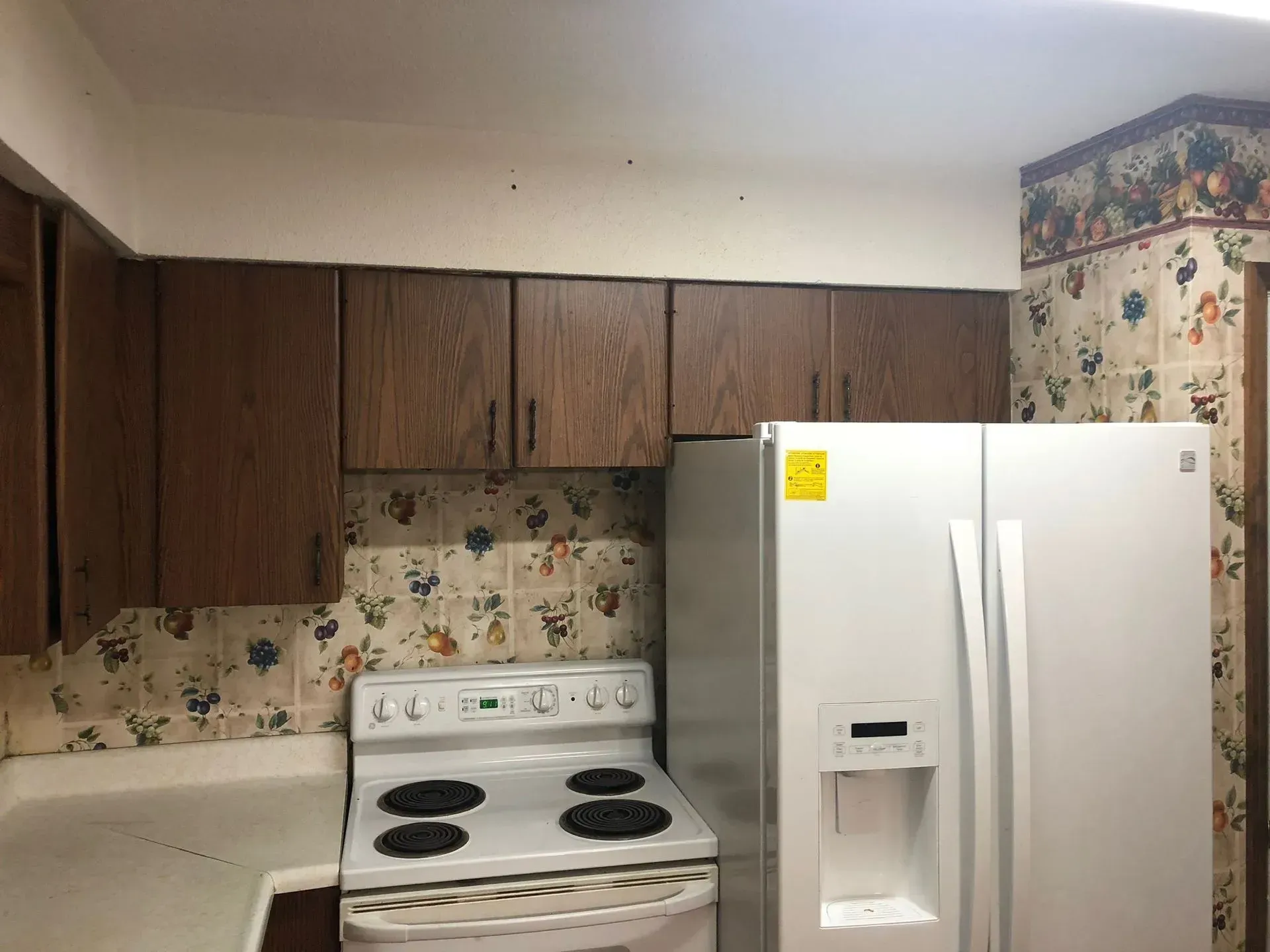 Kitchen with white appliances, brown cabinets, floral wallpaper, and a white countertop.