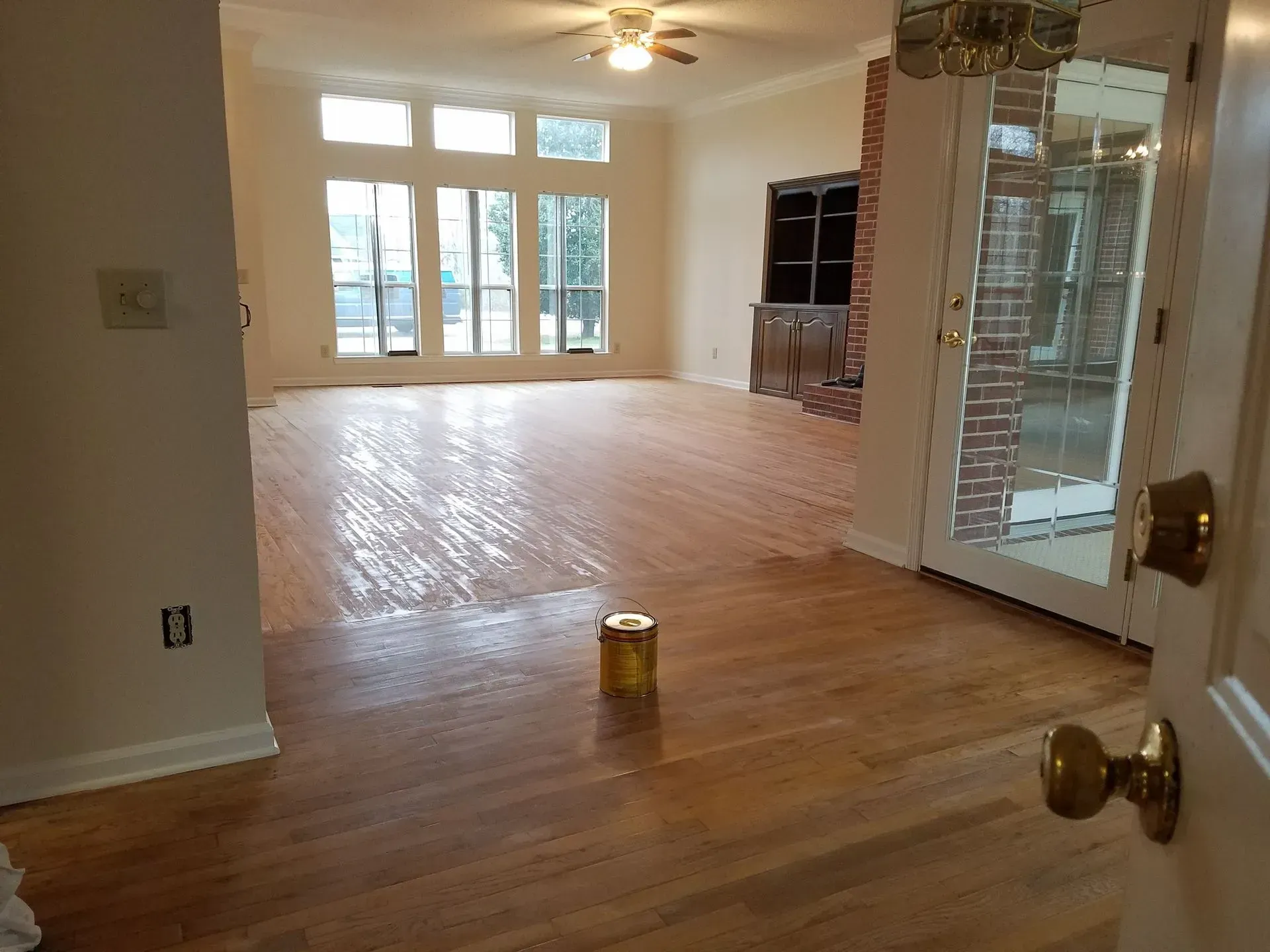 Empty living room with refinished wood floors and large windows; a door is in the foreground.