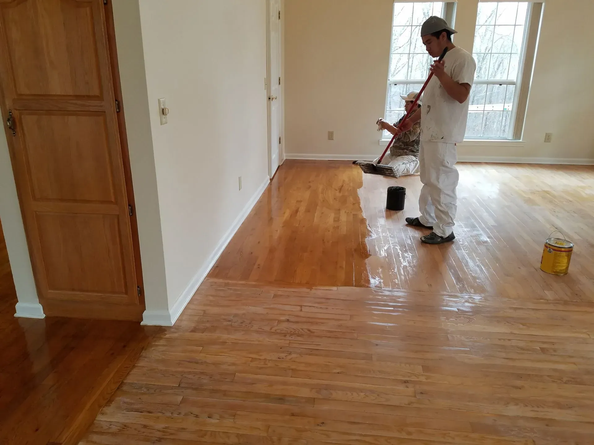 Two people applying finish to a hardwood floor. One applies with a long handle, another is in the background.