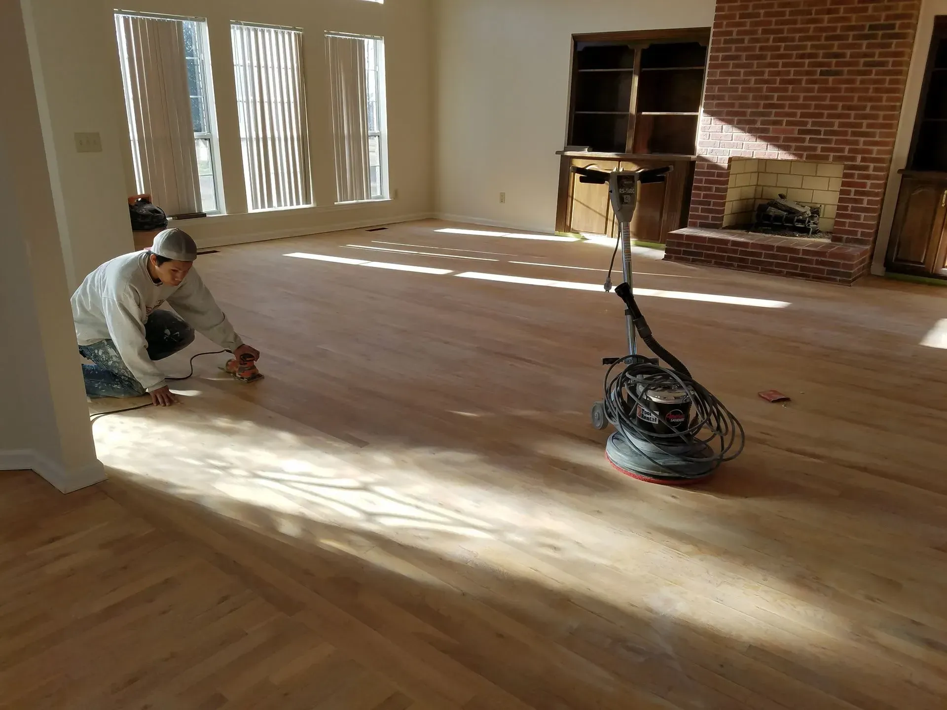 Person sanding a light-colored wood floor in a room with a brick fireplace and built-in cabinets.