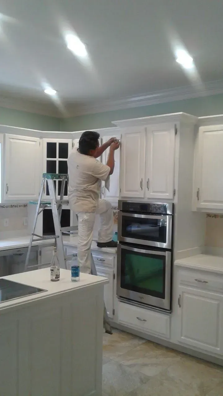 A person on a ladder is working on white kitchen cabinets, with a double oven and a kitchen island.