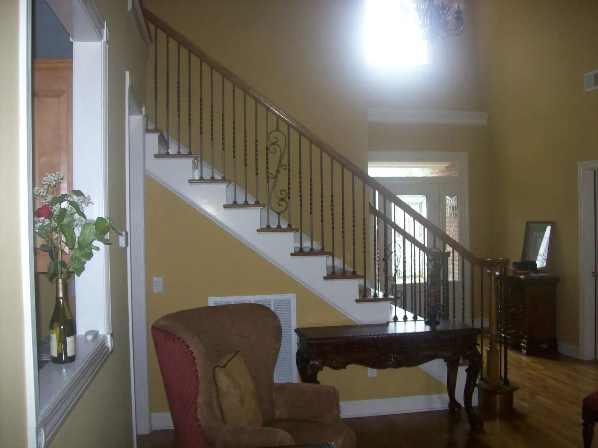 Staircase with dark metal railing, yellow walls, wooden console, brown chair.