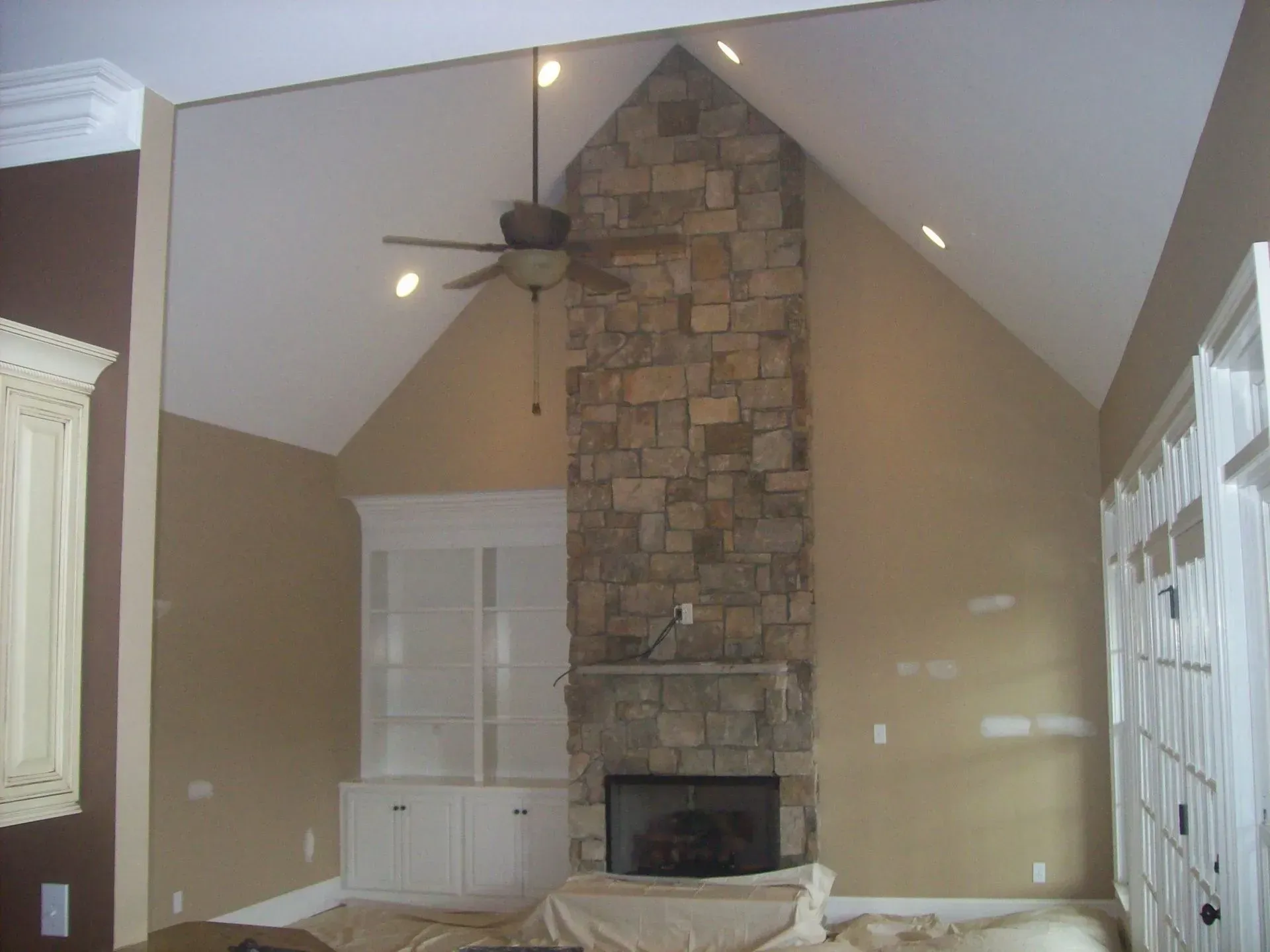 A living room with a stone fireplace, tan walls, high ceiling, and built-in cabinet.