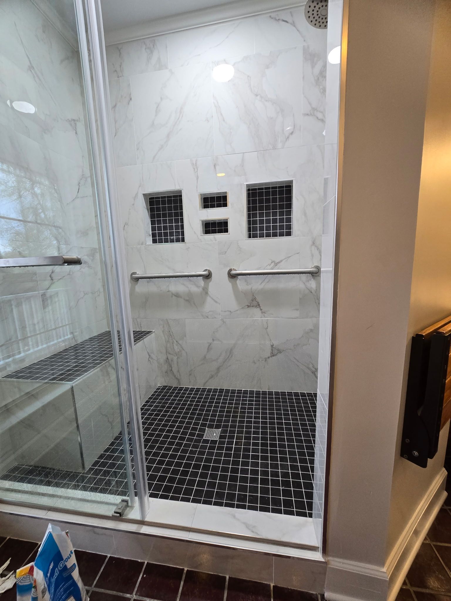 Modern shower with glass door, marble walls, chrome grab bars, and black bath mat